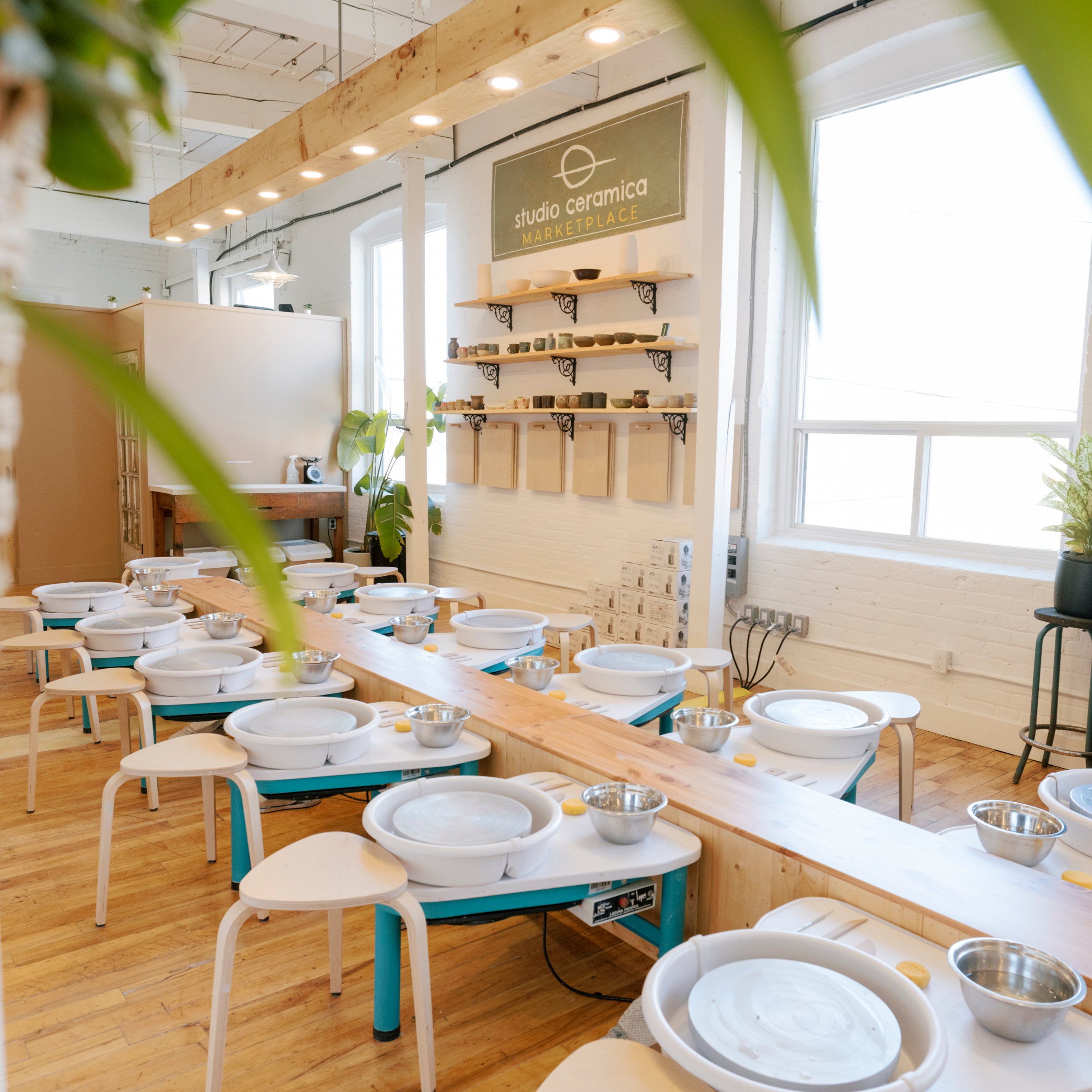 A pottery class studio with multiple wheel setups arranged along a long wooden table, bright natural light from large windows, and a visible 'elite ceramics' sign on the wall.