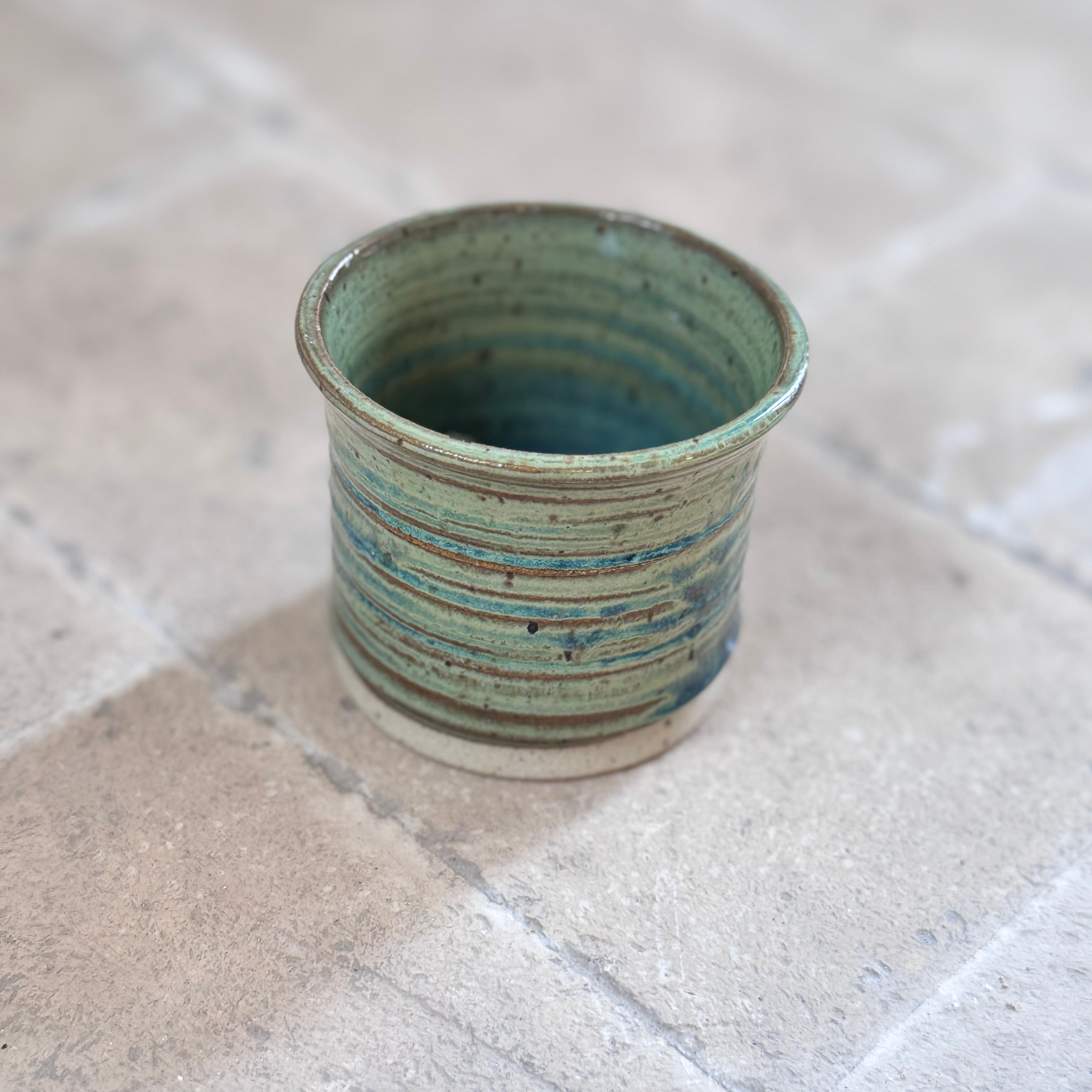 A handmade ceramic cup with a textured, ribbed exterior in a sage green and blue glaze, resting on a light beige stone tile surface.
