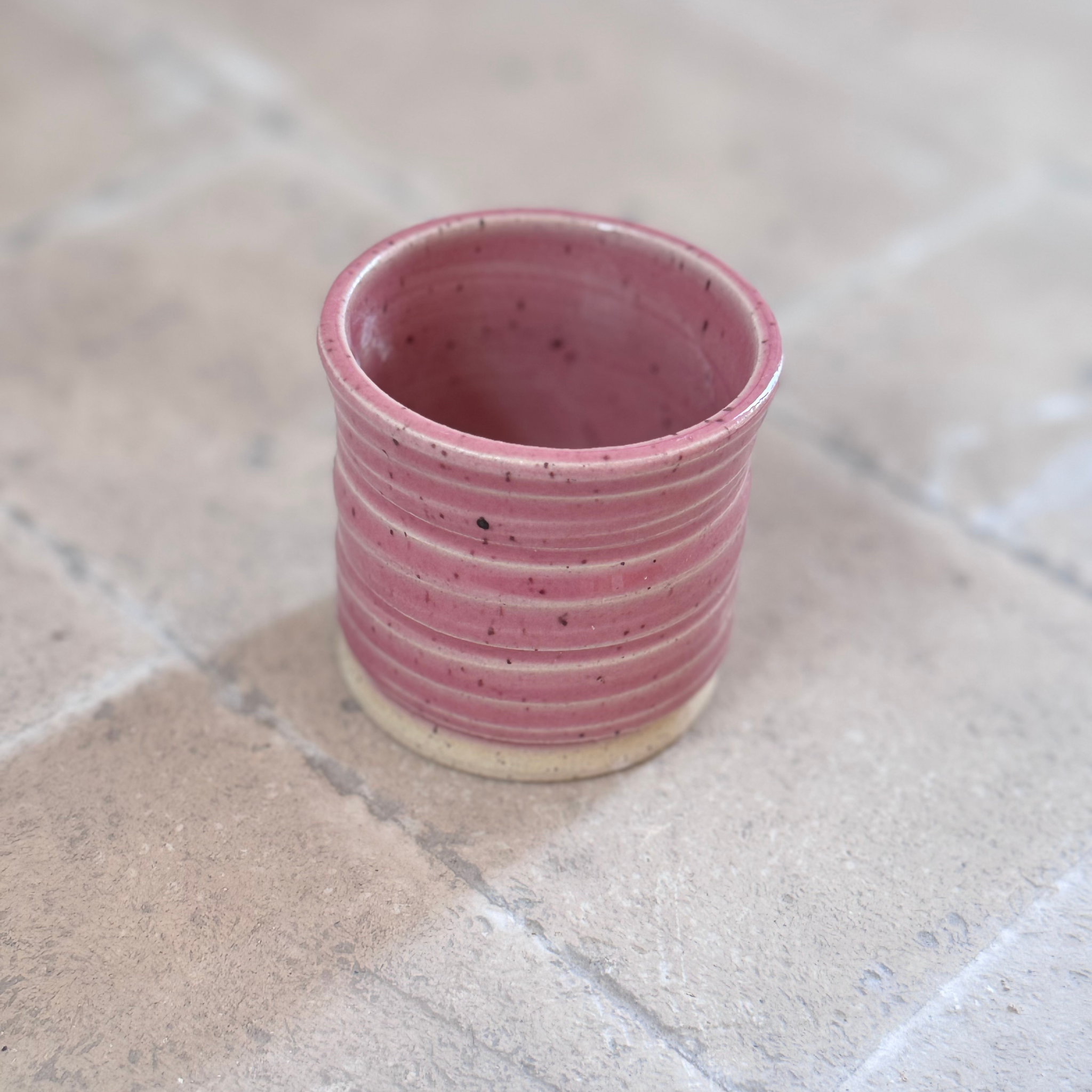 A small handmade ceramic cup with a dusty pink glaze and horizontal ridges, resting on a light beige stone tile surface.