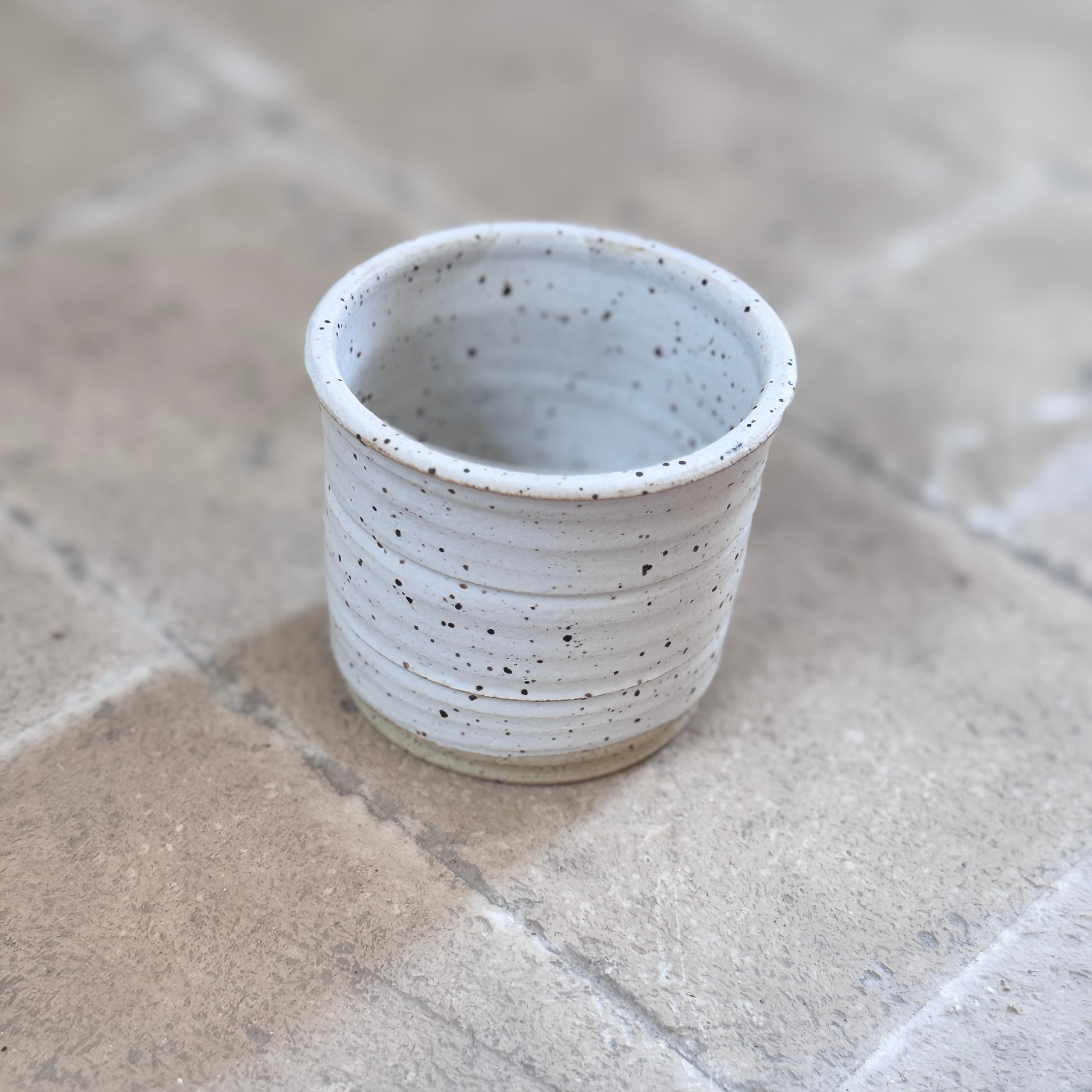 A small handmade ceramic cup with a speckled off-white glaze, standing on a stone tile surface.