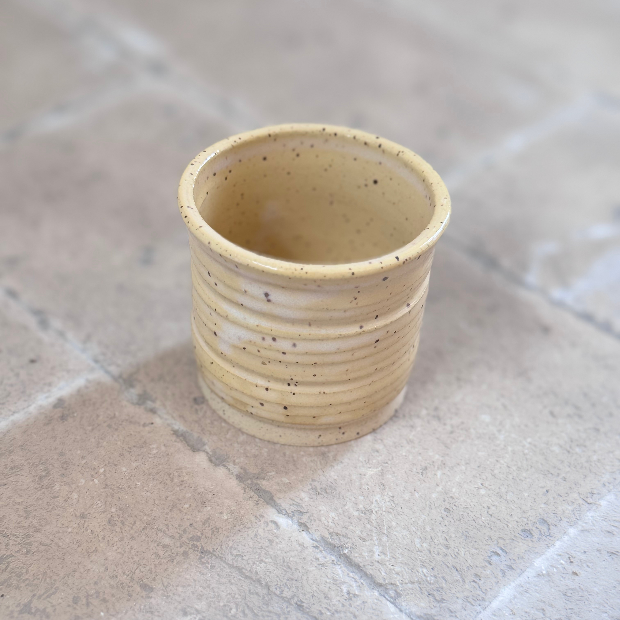 A small handmade ceramic cup with a flared rim, featuring horizontal ridges and speckled beige and cream glaze, set on a stone tile surface.