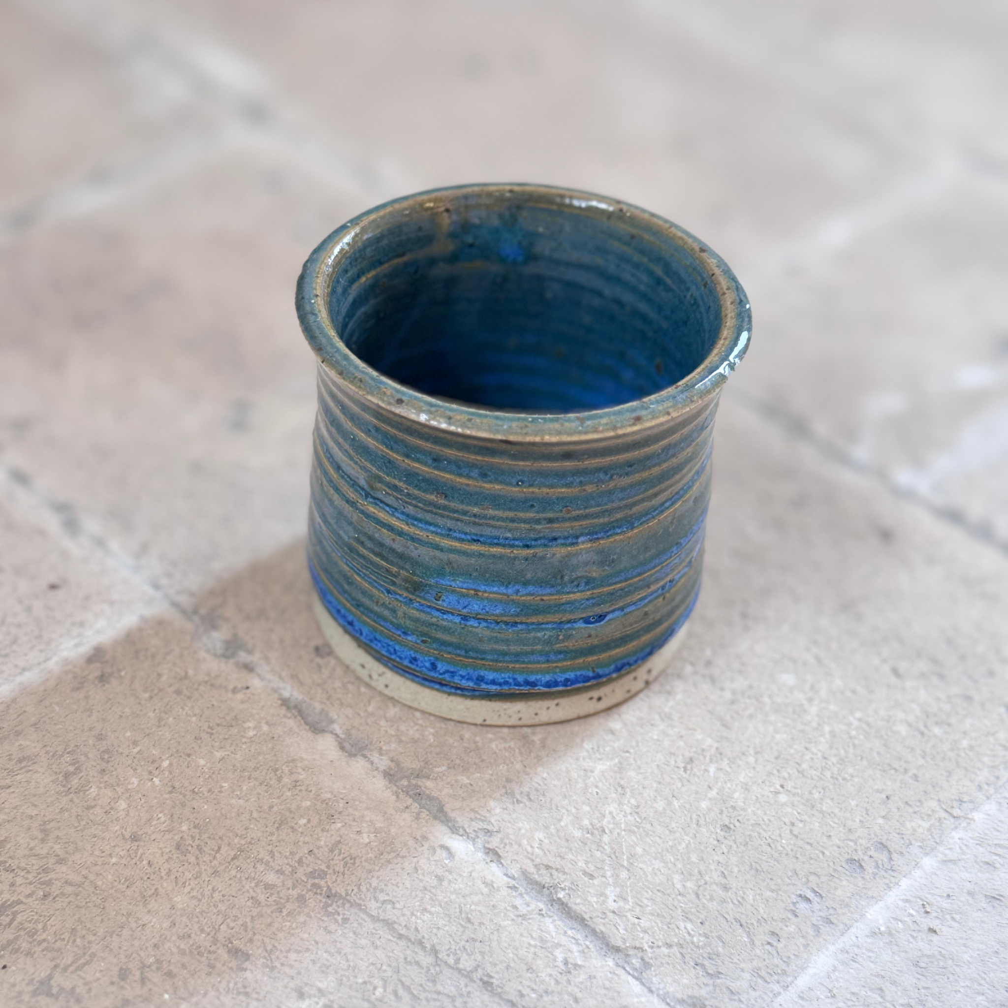 A small handmade ceramic cup with a blue and green striped glaze, standing on a light beige stone tile surface.