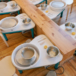 A pottery class setup with multiple white and teal wheel stations arranged on a wooden floor, each with a metal bowl, small yellow wheel key, and a spinning clay wheel.