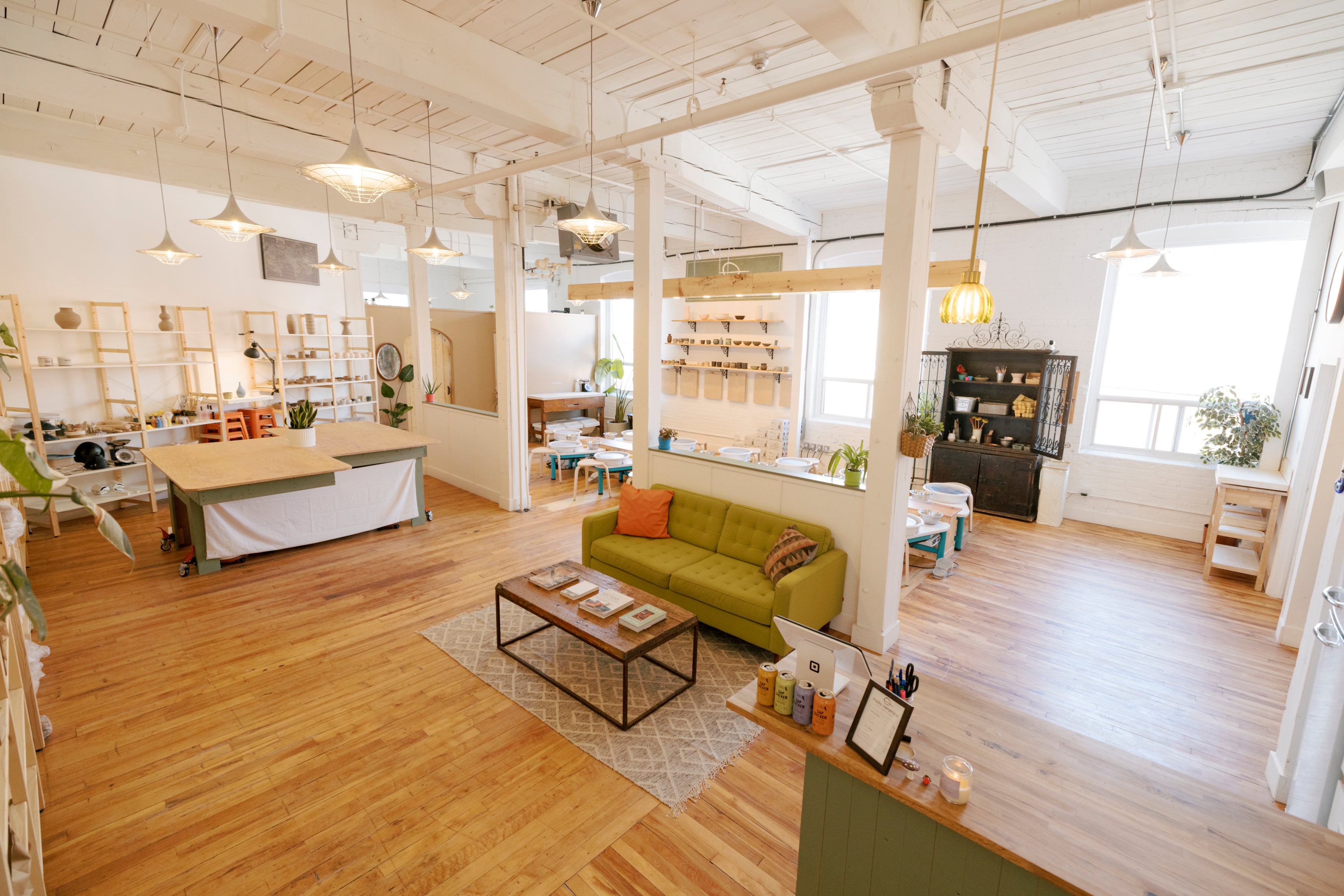 A bright, modern loft-style room with exposed wood ceiling beams, white columns, and light hardwood floors. The space includes a green sofa with orange accent pillows, a small black coffee table, shelving units, hanging pendant lights, and a reception desk in the foreground.