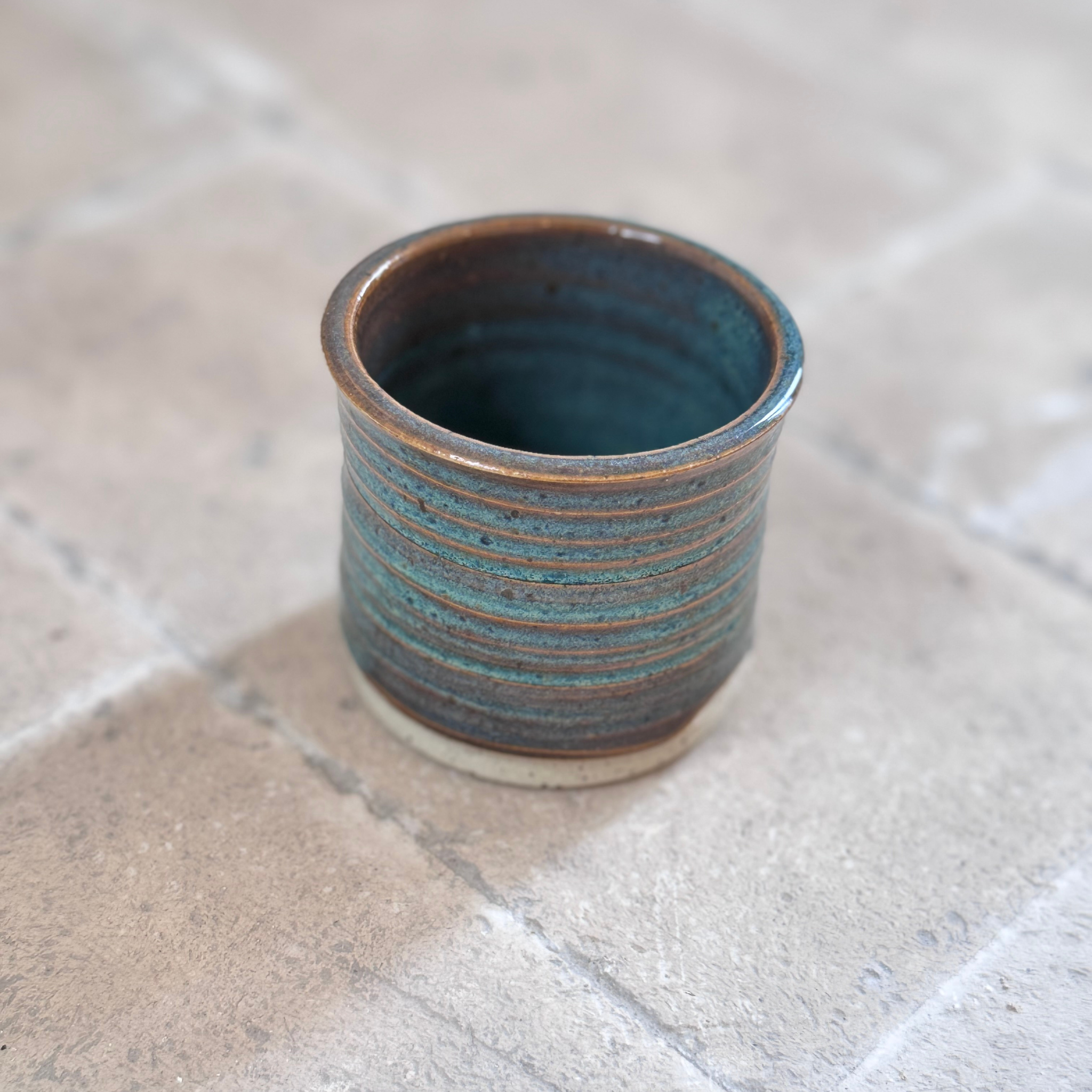 A small handmade ceramic cup with a textured, ribbed exterior and a glazed interior showing shades of teal and brown, set on a light stone tile surface.