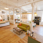 A bright, modern loft-style room with exposed wood ceiling beams, white columns, and light hardwood floors. The space includes a green sofa with orange accent pillows, a small black coffee table, shelving units, hanging pendant lights, and a reception desk in the foreground.