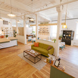A bright, modern loft-style room with exposed wood ceiling beams, white columns, and light hardwood floors. The space includes a green sofa with orange accent pillows, a small black coffee table, shelving units, hanging pendant lights, and a reception desk in the foreground.