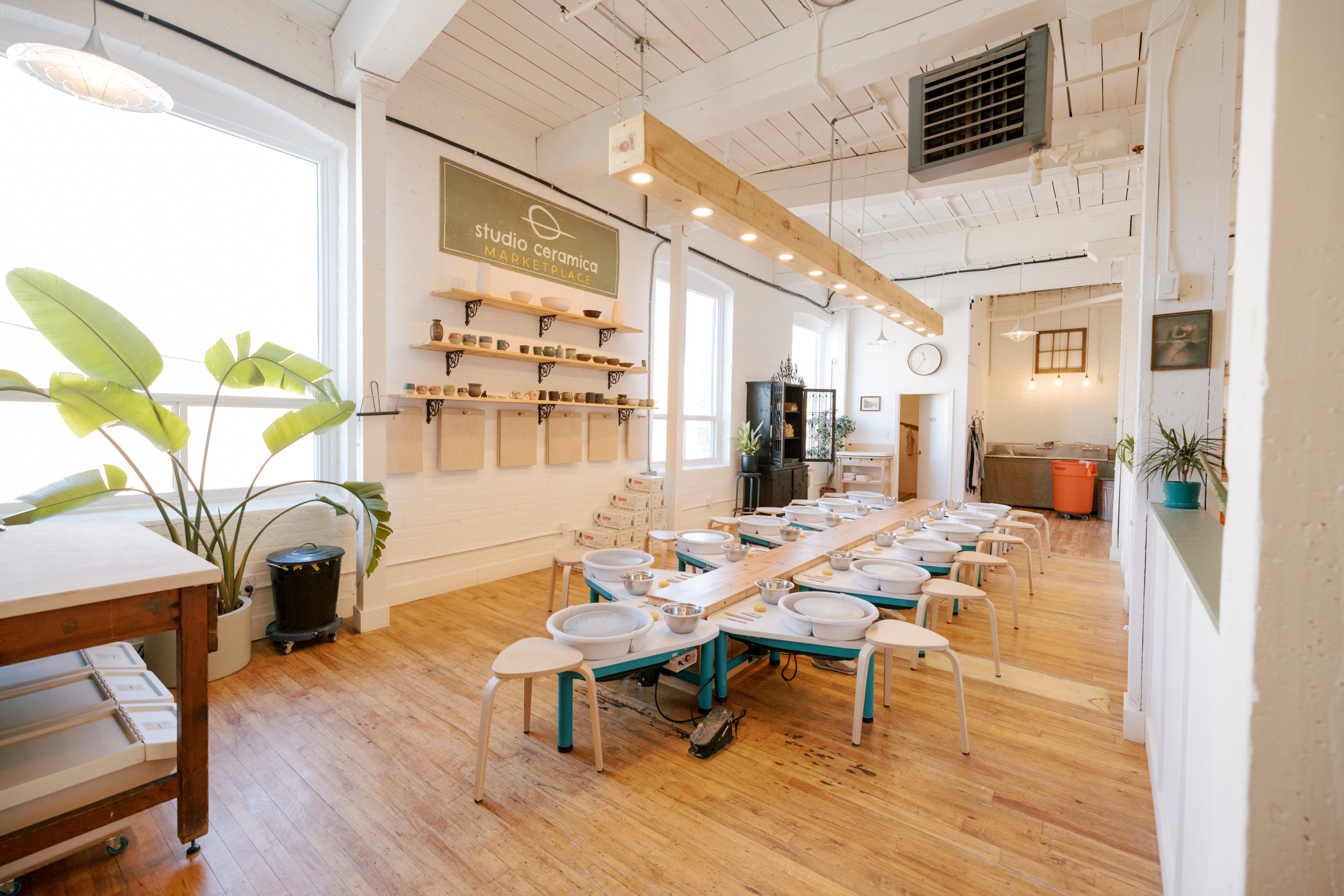 A bright, modern ceramic studio with wooden floors, white walls, and a long table set with multiple white ceramic bowls and small chairs. A sign on the wall reads 'studio ceramics', and large potted plants are placed near the windows.