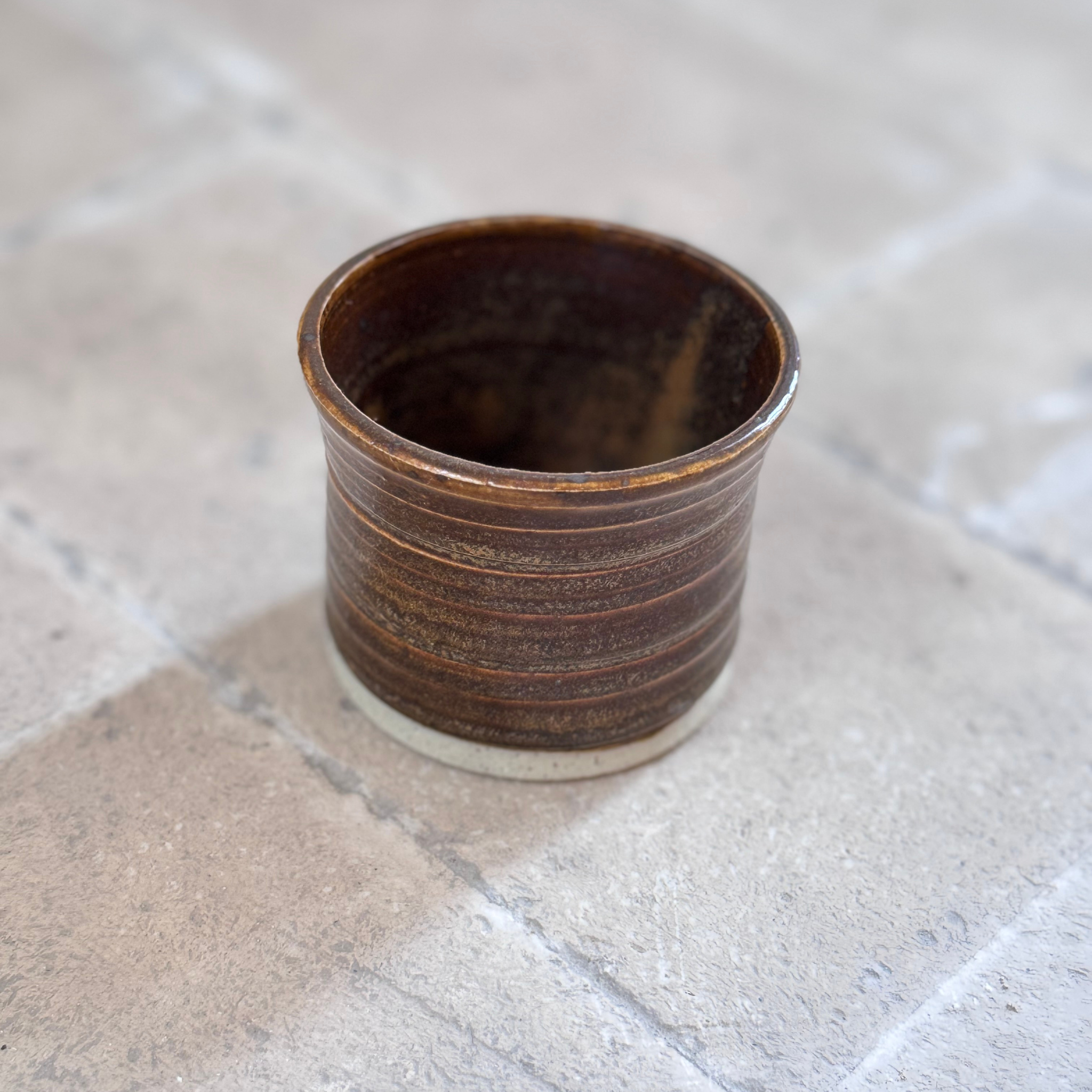 A small handmade ceramic cup with a rustic dark brown and amber-brown glaze, resting on a light gray stone tile surface.