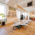 A bright, modern ceramic studio with wooden floors, white walls, and a long table set with multiple white ceramic bowls and small chairs. A sign on the wall reads 'studio ceramica', and large potted plants are placed near the windows.