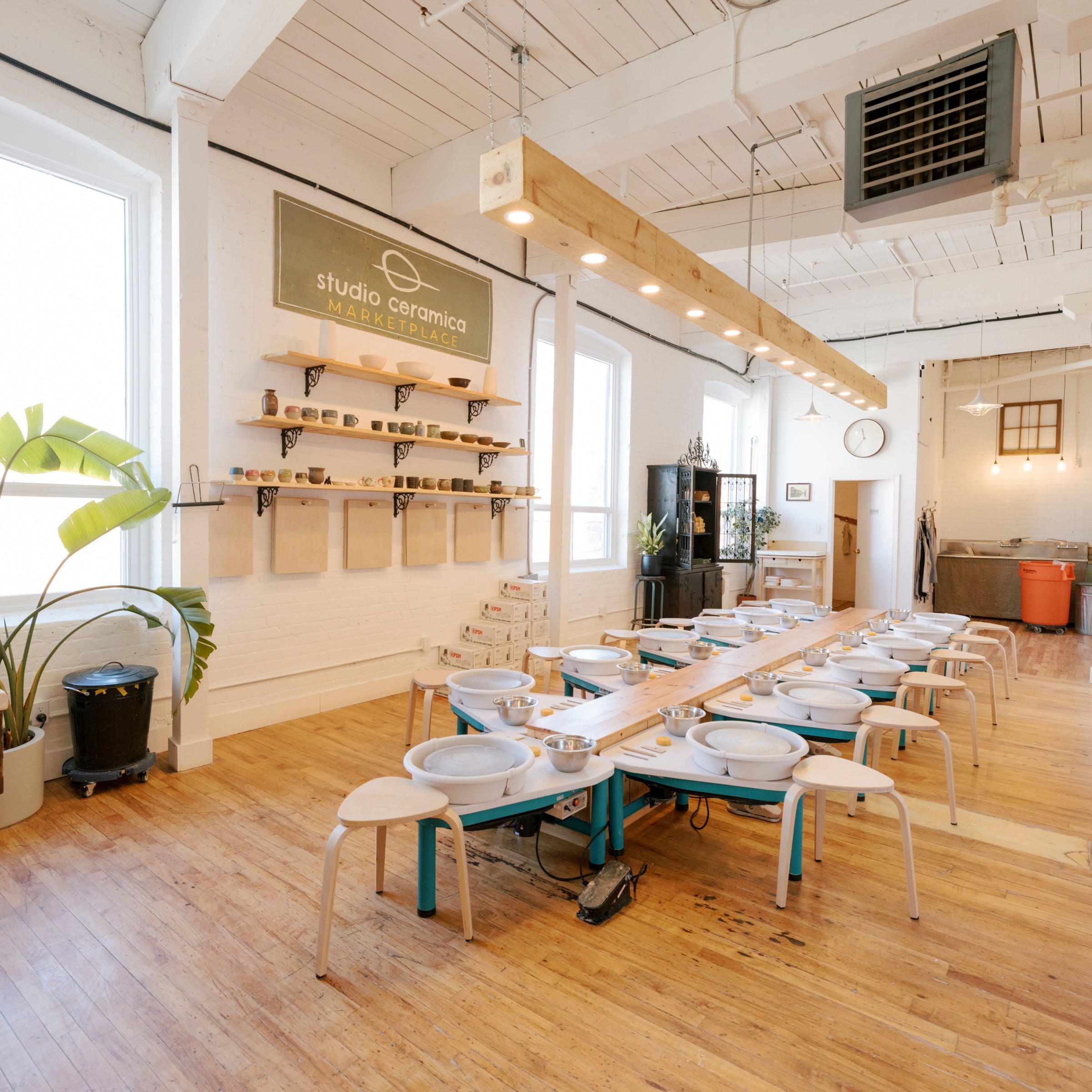 A bright, modern ceramic studio with wooden floors, white walls, and a long table set with multiple white ceramic bowls and small chairs. A sign on the wall reads 'studio ceramica', and large potted plants are placed near the windows.