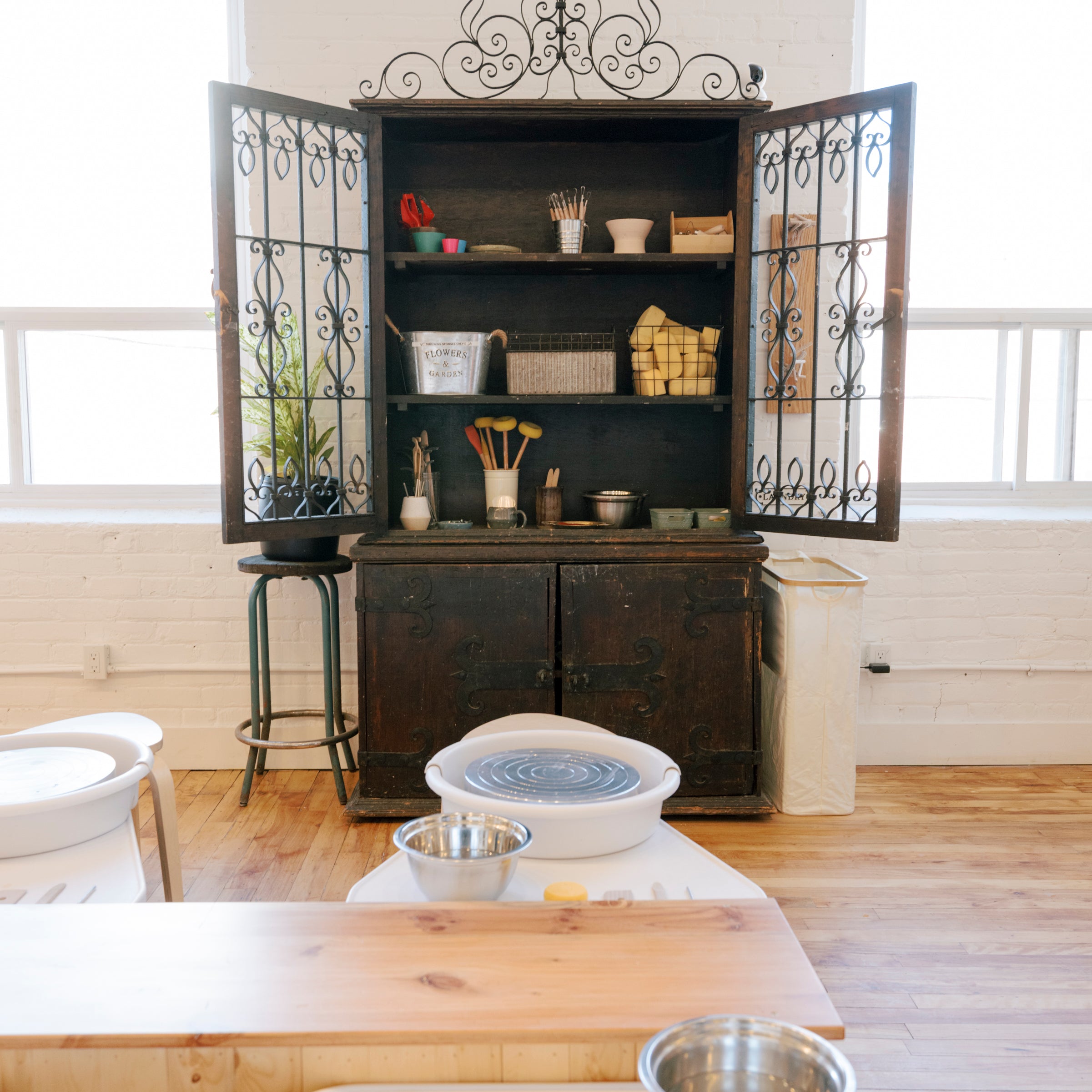 A rustic dark wood hutch cabinet with open doors and decorative ironwork, styled with pottery wheels, metal bowls, and baskets in a bright room with wood floors and white walls.
