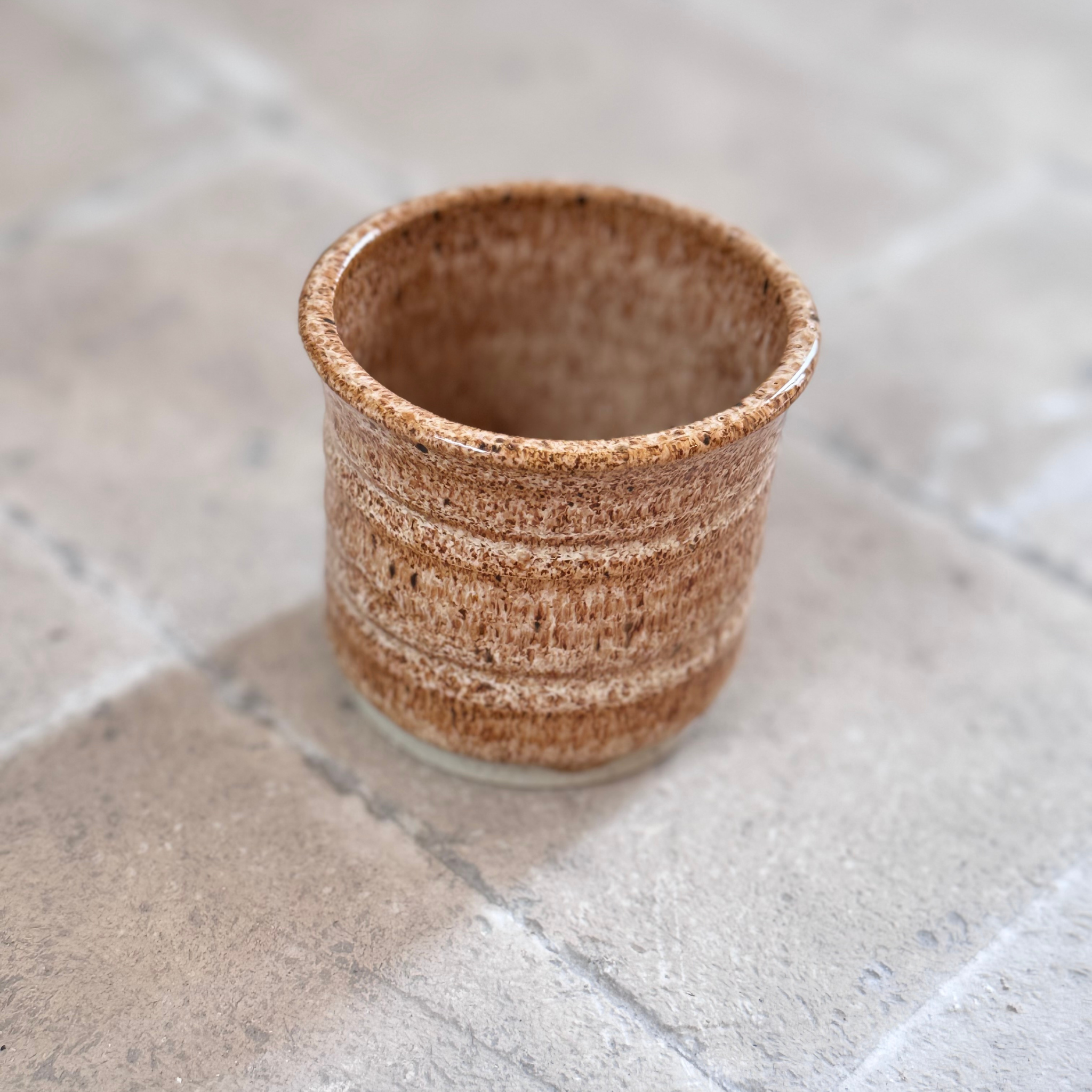 A small handmade ceramic cup with a textured, speckled brown and beige glaze, shown on a light gray stone tile surface.