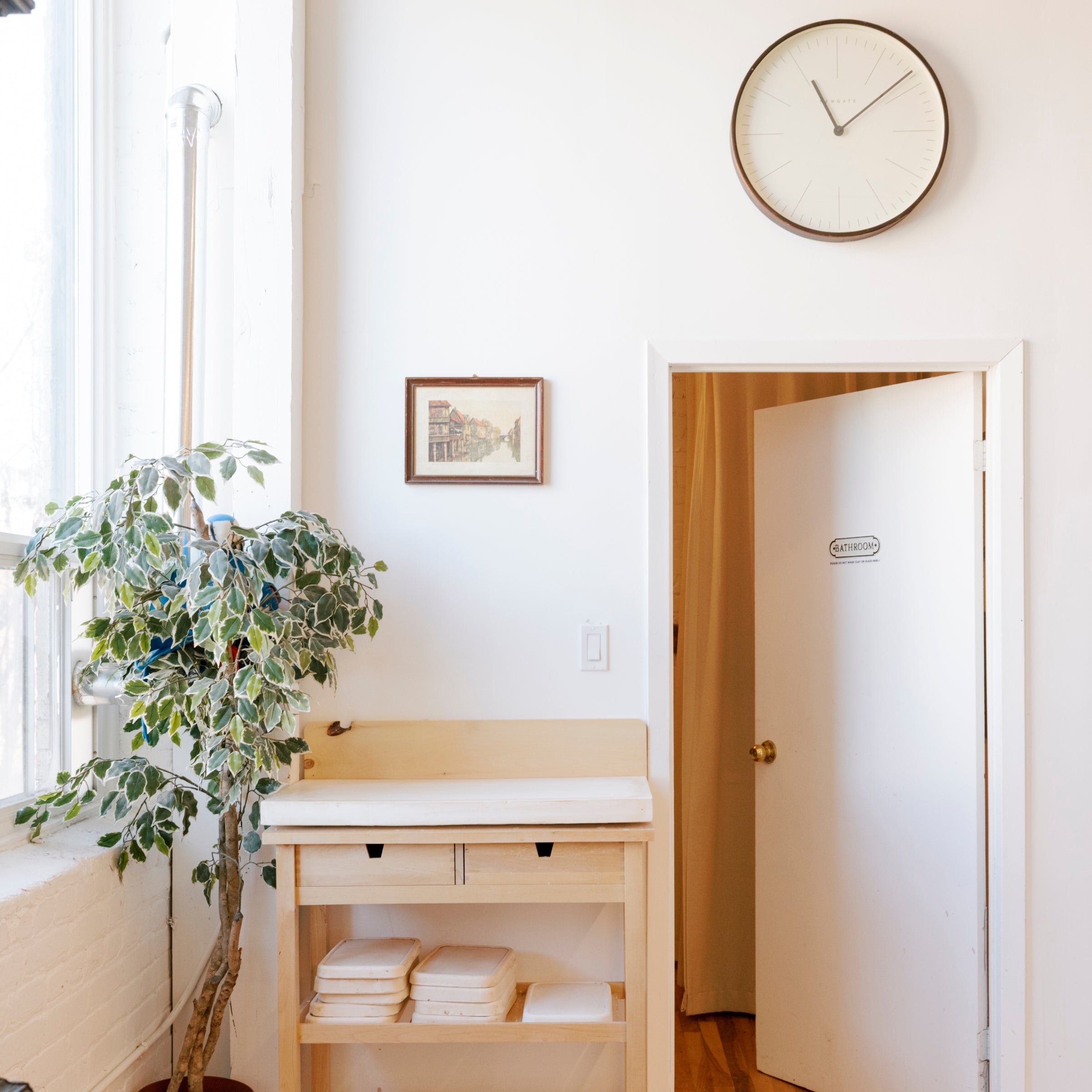 A light wood changing table with a white top sits in a bright, minimalist room with hardwood floors, a potted plant, a wall clock, and an open door.