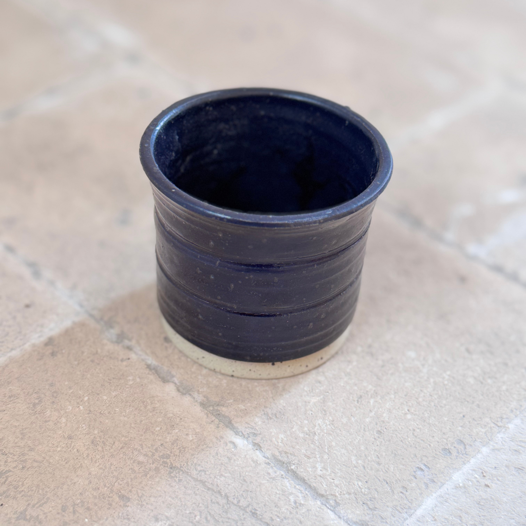 A small handmade ceramic cup with a dark navy blue glazed finish and a natural beige clay base, set on a light beige tiled surface.