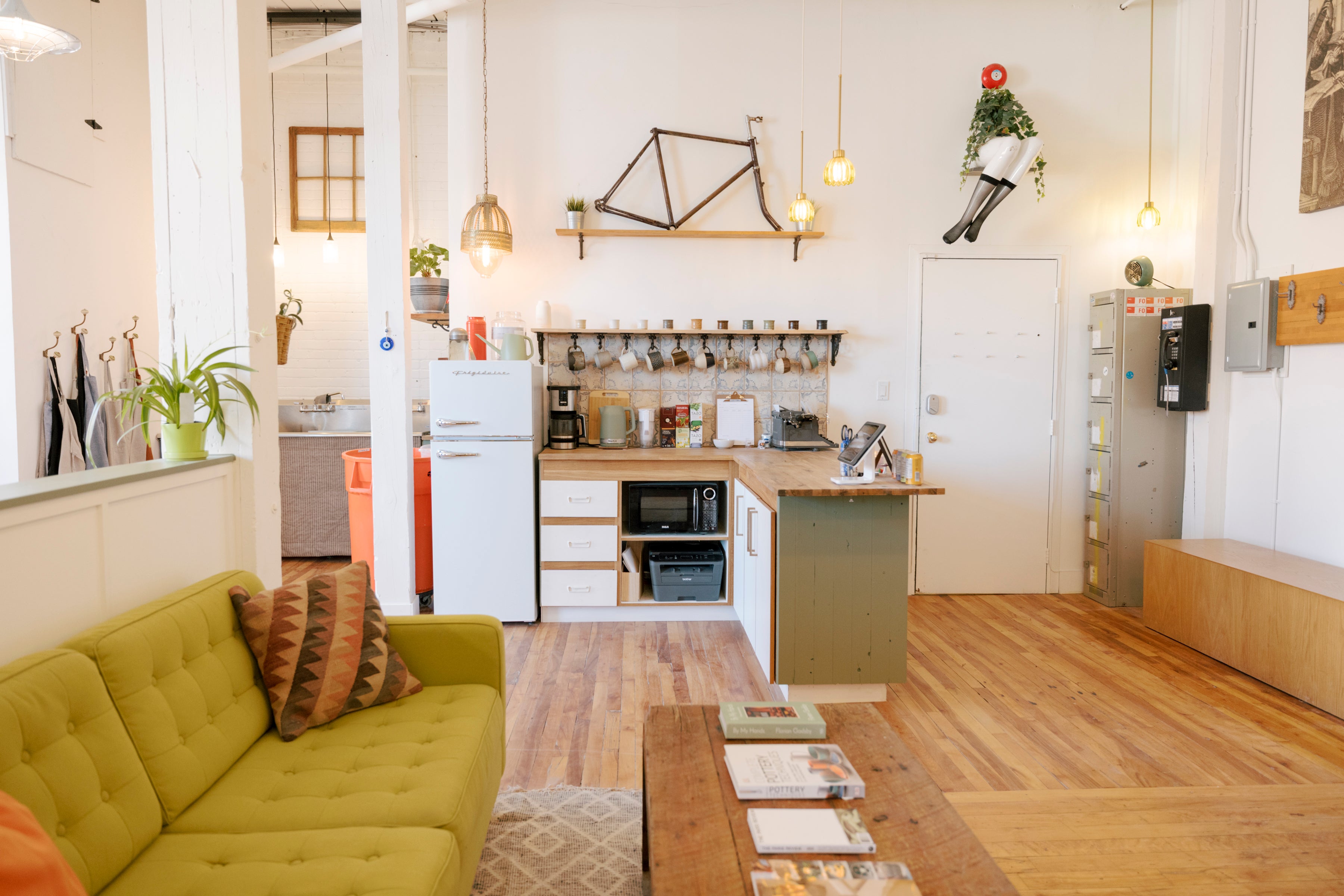A bright, modern open-plan living and kitchen space with a mustard yellow tufted sofa in the foreground, a wooden coffee table, and a compact kitchen area with white appliances and wood countertops in the background.