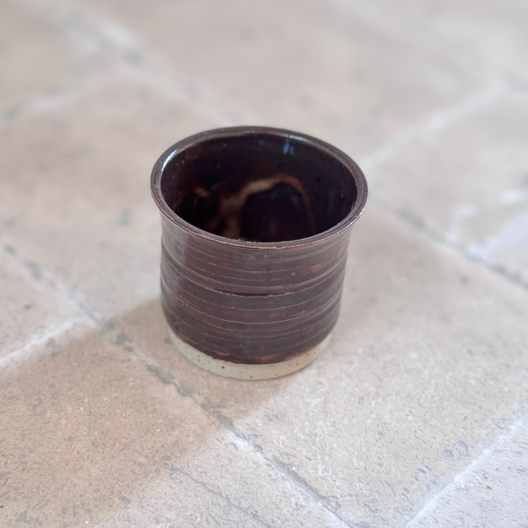 A small handmade ceramic cup with a dark brown glazed finish and horizontal ridges, resting on a light beige stone tile surface.