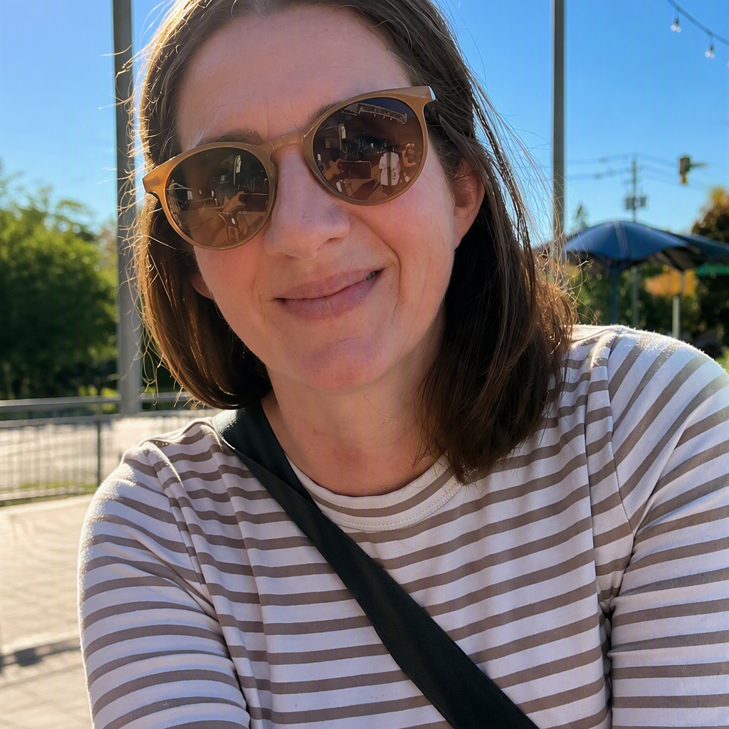 A woman with brown hair wearing brown cat-eye sunglasses and a beige and white striped t-shirt, with a black strap across her chest, photographed outdoors on a sunny day.