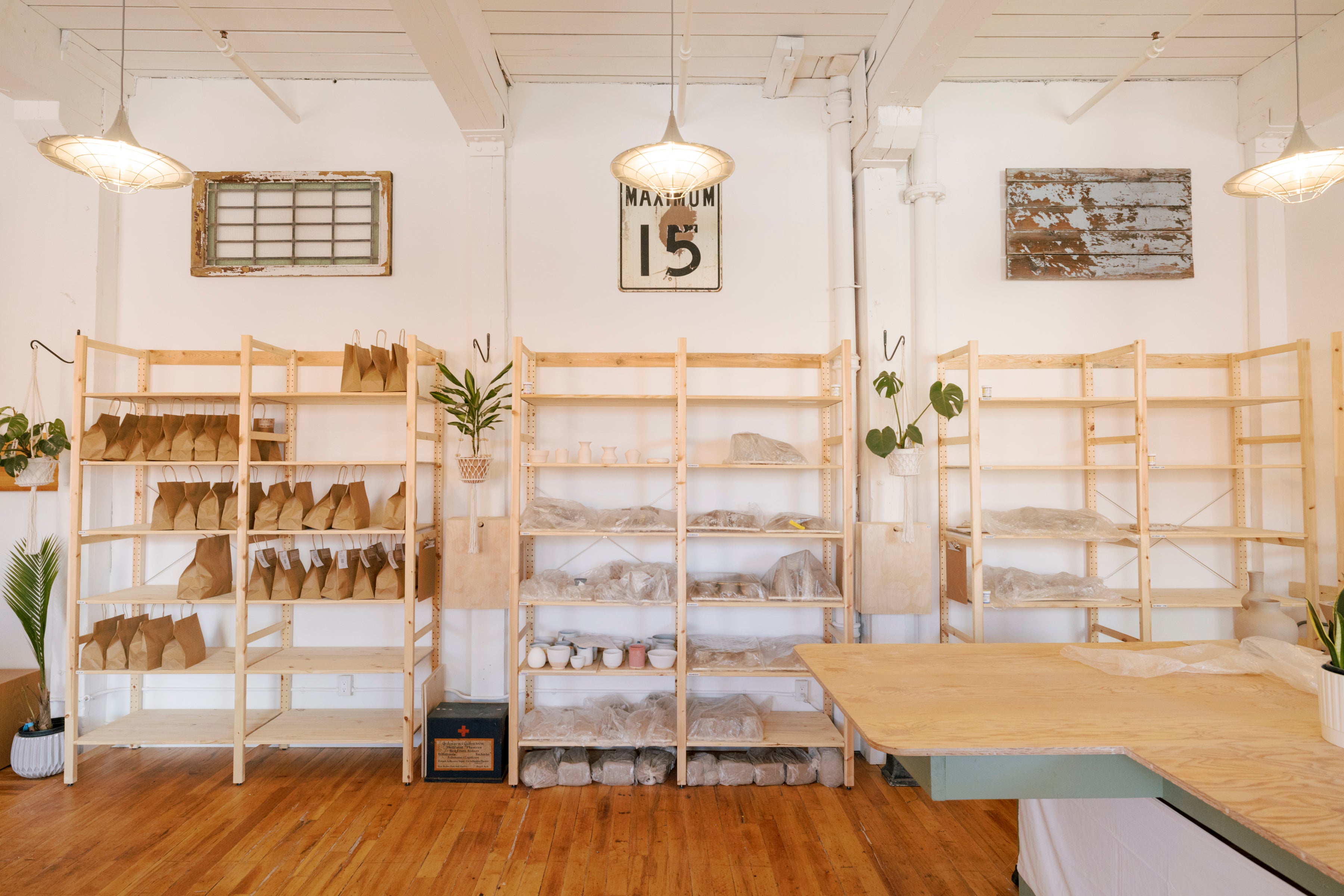 A bright, airy craft room with light wood shelving units filled with leather and fabric materials, a wooden worktable in the foreground, and warm wood floors. A framed 'MAYUN 15' sign hangs on the white wall.