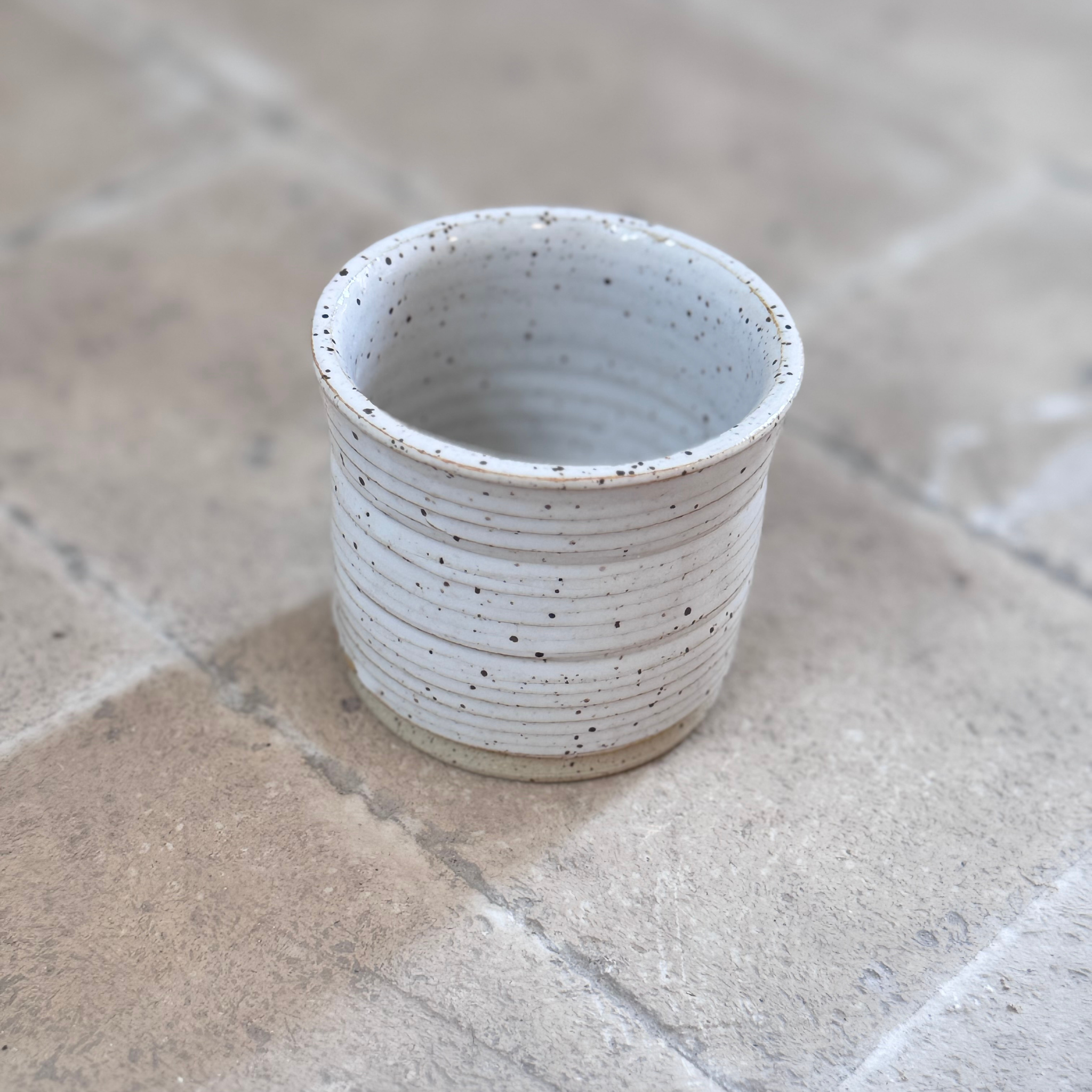 A small handmade ceramic cup with a speckled off-white glaze and horizontal ridges, sitting on a stone tile surface.