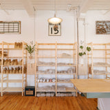 A bright, airy craft room with light wood shelving units filled with leather and fabric materials, a wooden worktable in the foreground, and warm wood floors. A framed 'MAYUN 15' sign hangs on the white wall.
