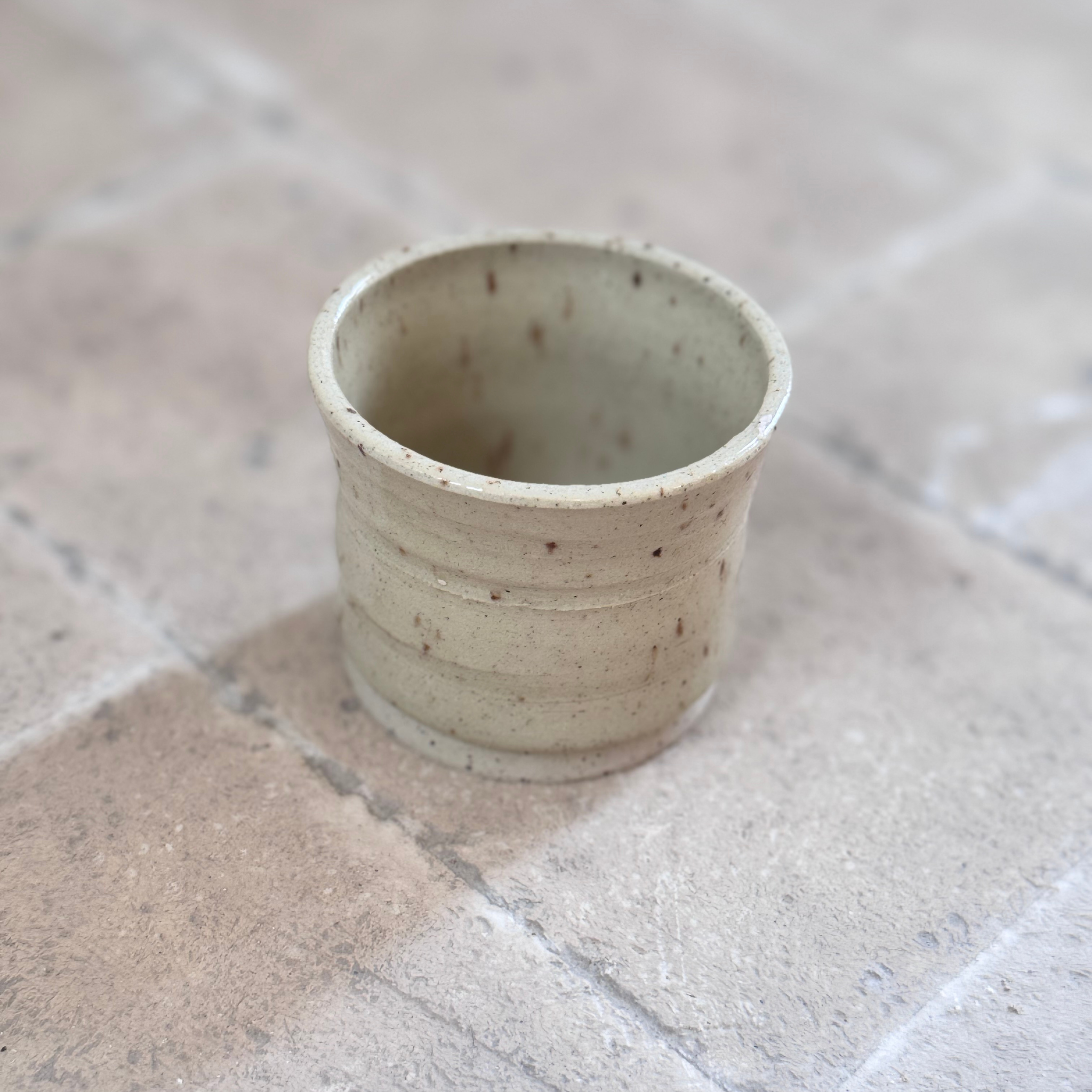 A small handmade ceramic cup with a speckled beige and off-white glaze, standing on a stone-tiled surface.