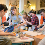 A group of people participating in a pottery class, working at wheel-mounted potters' wheels and shaping clay in a bright, warm indoor studio setting.