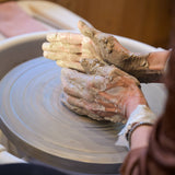 Close-up of a person's hands covered in wet clay shaping a piece on a spinning pottery wheel, with the wheel and a warm wooden background visible.