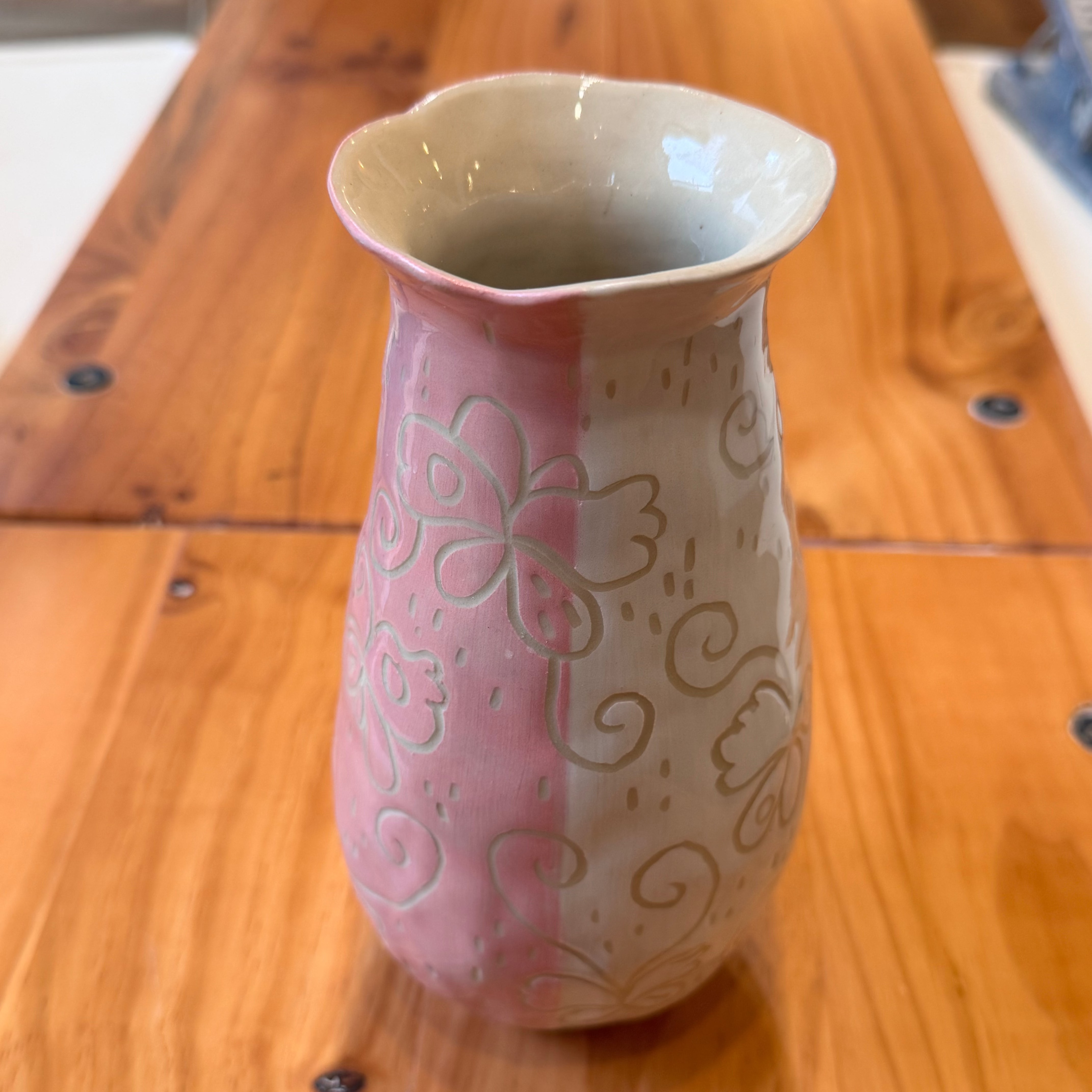 A small ceramic vase with a split pink and beige design, featuring embossed sgraffito floral and scroll patterns, sitting on a wooden table.