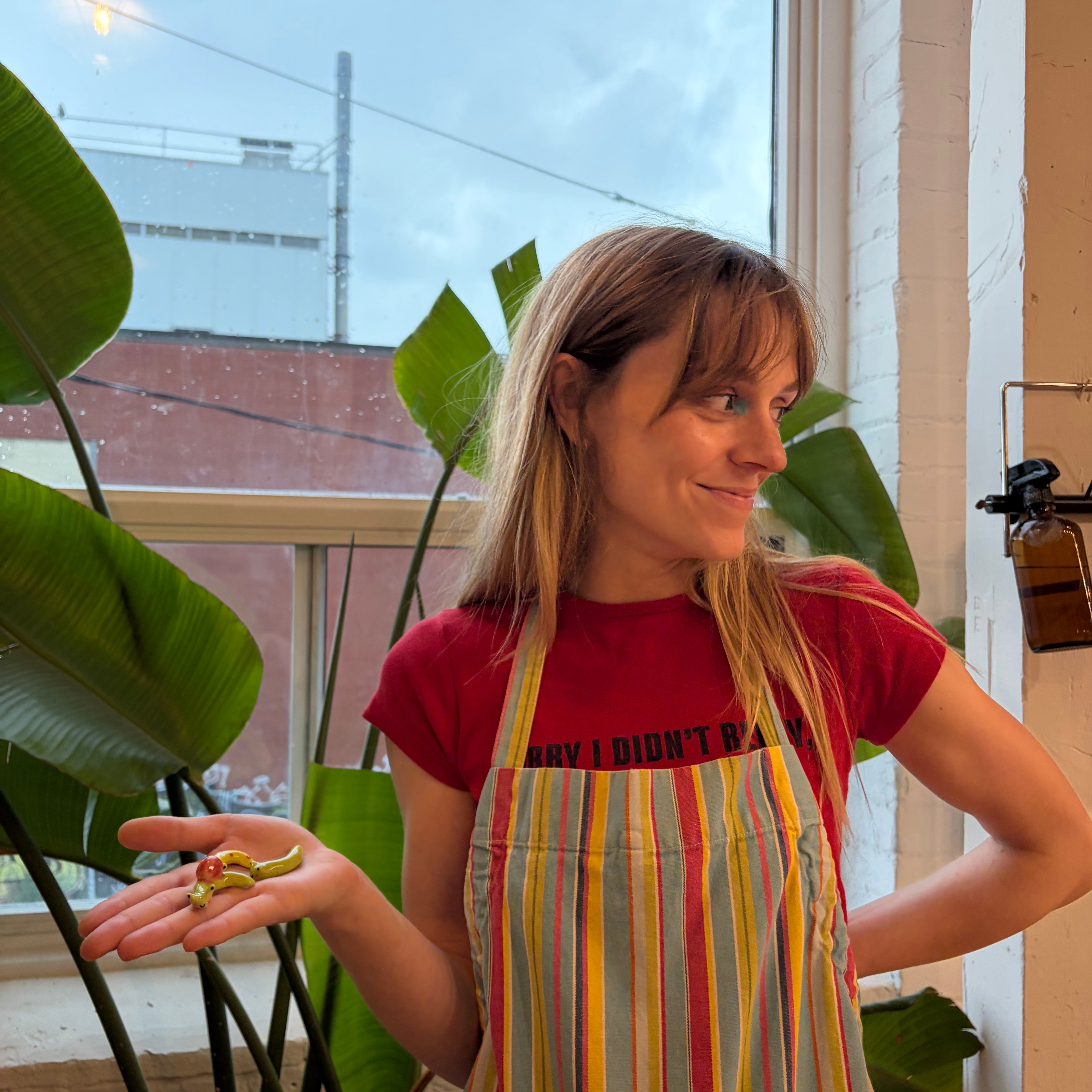 A woman wearing a red graphic t-shirt and a colorful vertically striped apron stands in a bright room with large green plants and a window behind her.
