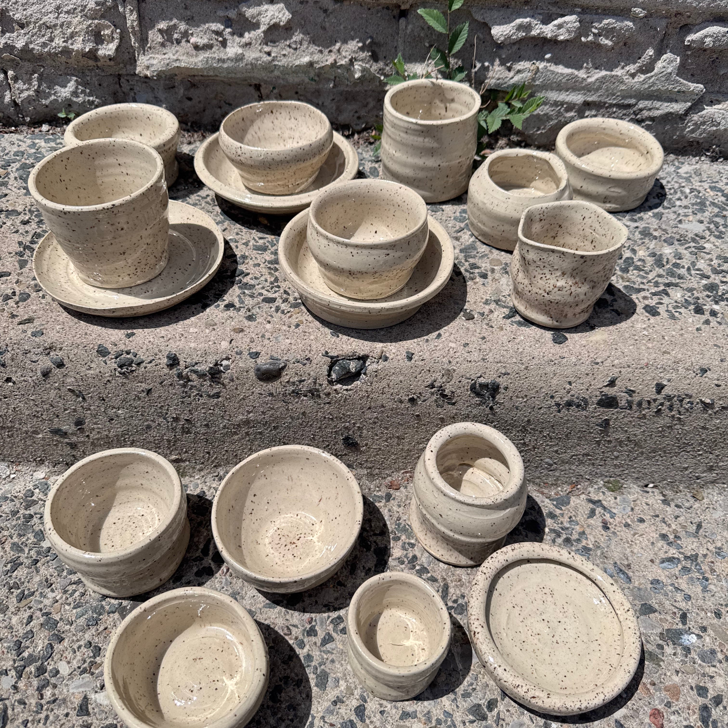 A set of handmade ceramic planters and small cups in a speckled beige stoneware finish, arranged on concrete steps against a stone wall.