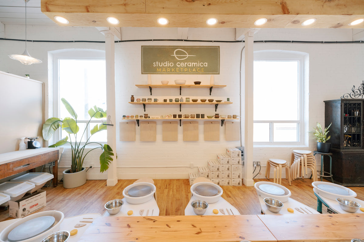 A pottery studio interior with multiple wheel kilns arranged around a large wooden table, shelves of ceramic pieces on the wall, and the sign 'studio ceramica pottery club' above. The room has white walls, wood floors, large windows, and potted plants.