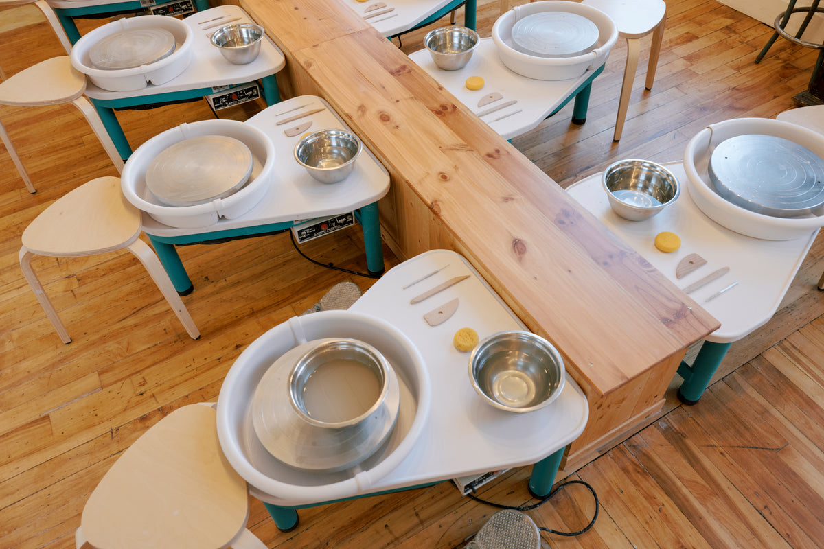 A pottery class setup with multiple white and teal wheel stations arranged on a wooden floor, each with a metal bowl, small yellow wheel key, and a spinning clay wheel.