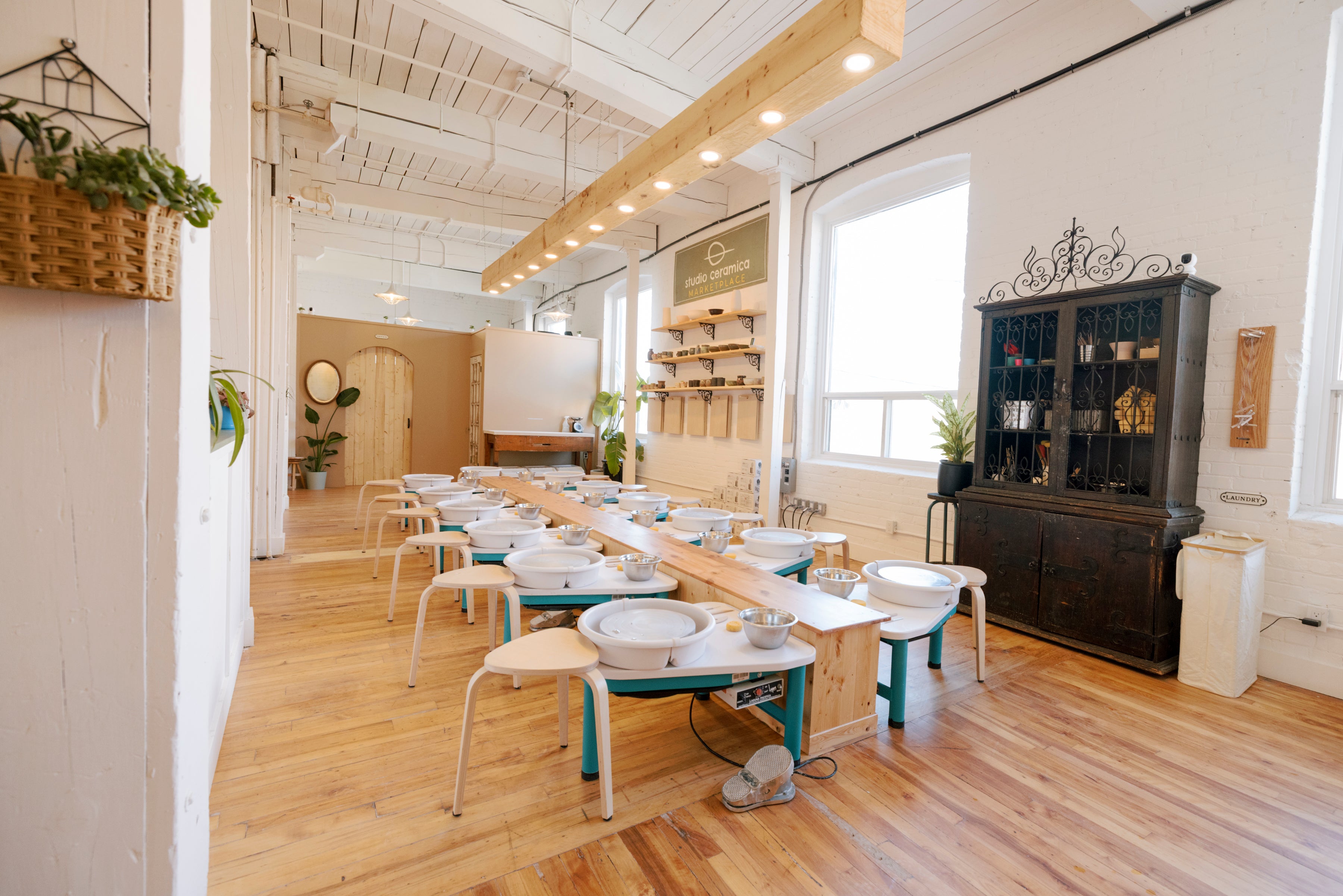 A bright, spacious pottery studio with hardwood floors, white walls, and large windows. Multiple pottery wheels with white bowls and clay pots are arranged in rows, with shelves, plants, and a dark wooden cabinet in the background.
