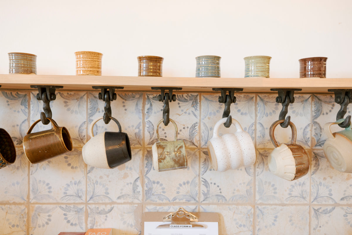 A shelf-mounted mug rack with black metal hooks holding various ceramic mugs and cups, with matching ceramic canisters arranged on the shelf above, set against a patterned tile backsplash.
