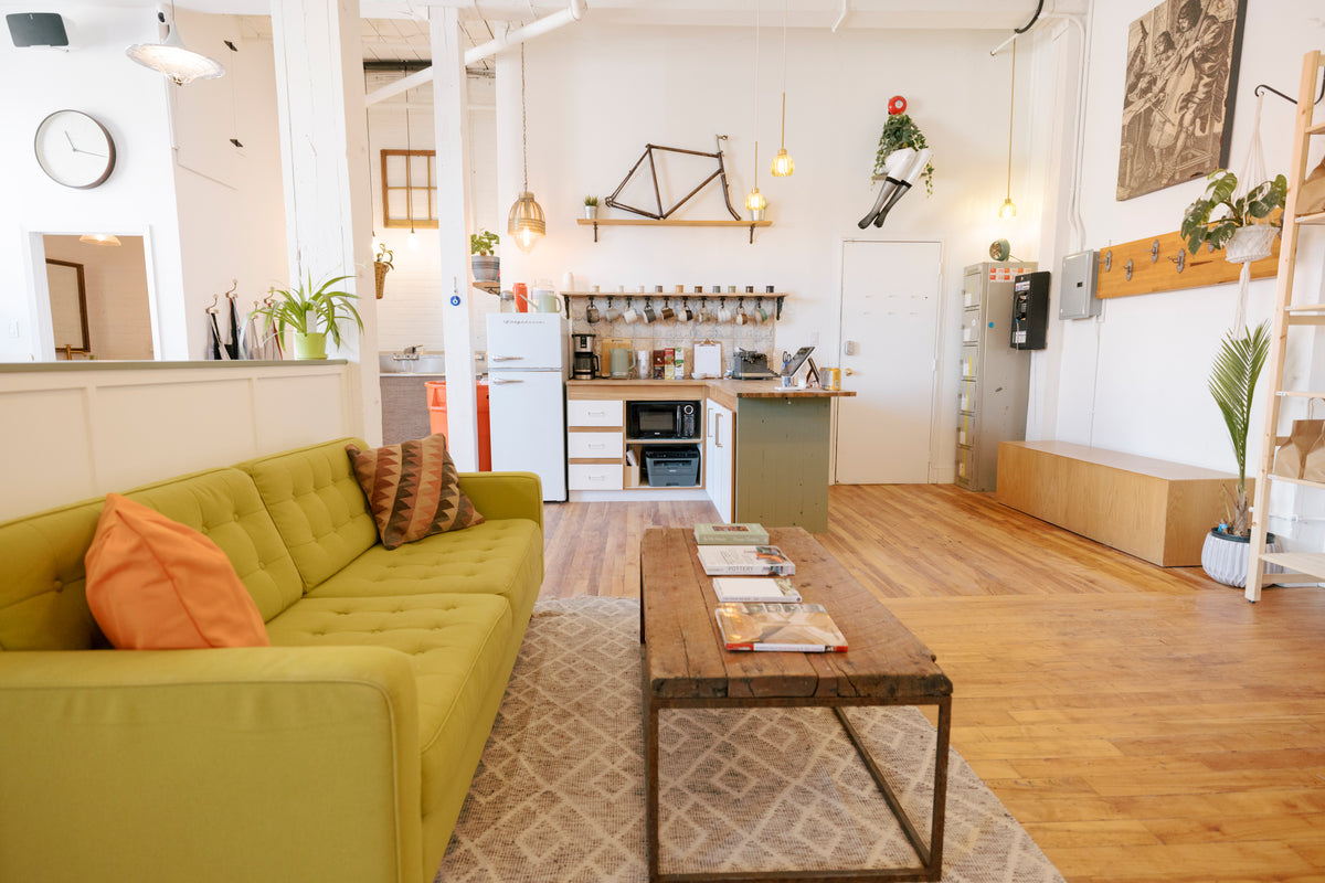 A bright, modern loft-style living room featuring a tufted lime green sofa with orange and brown accent pillows, a rustic wooden coffee table, and a light patterned rug. The open-plan space includes a small kitchen area in the background with white walls, wood floors, and hanging light fixtures.