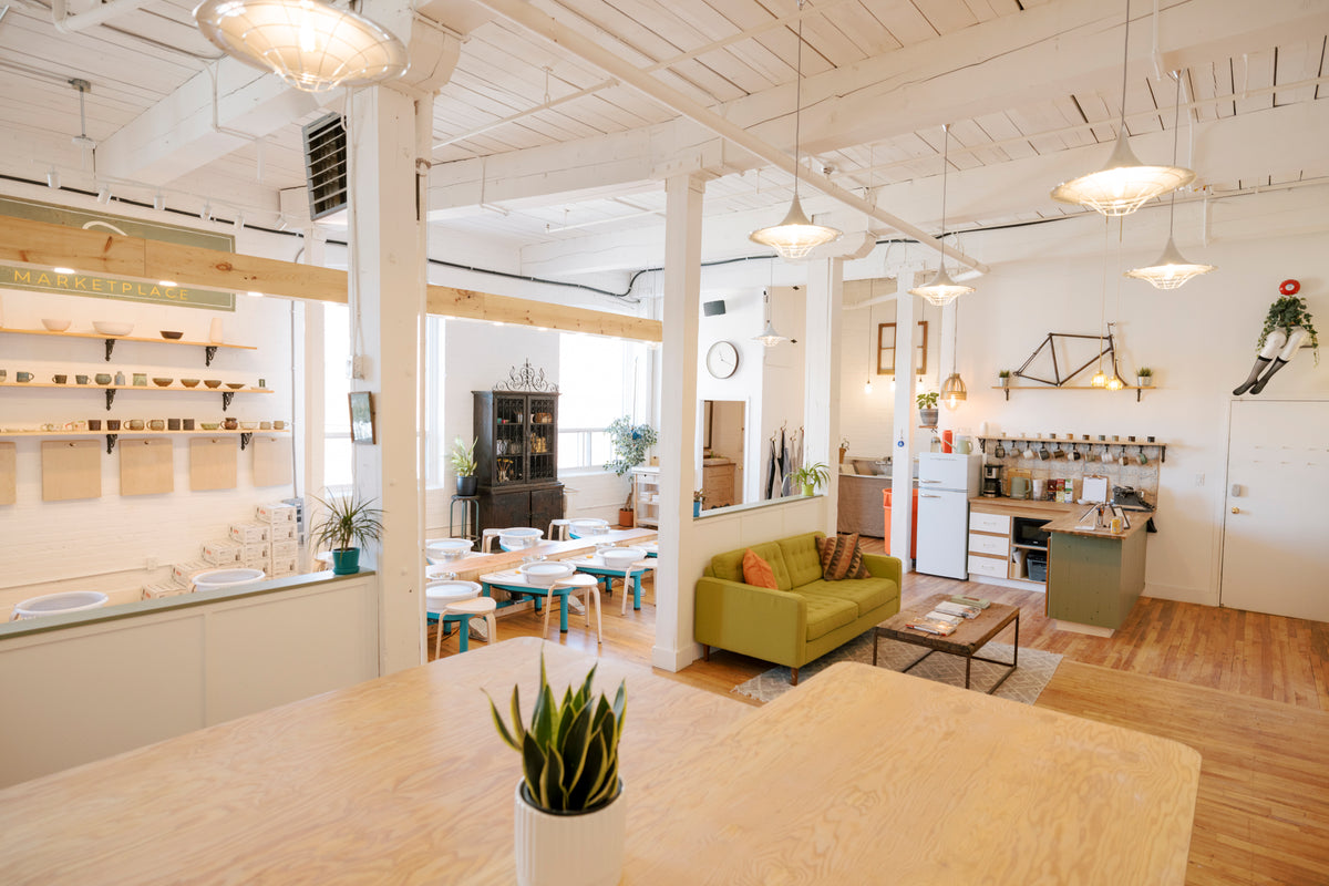 A bright, modern open-plan interior with a cafe or kitchen counter on the left, a green sofa in the living area, and pendant lights hanging from a white ceiling with exposed beams.