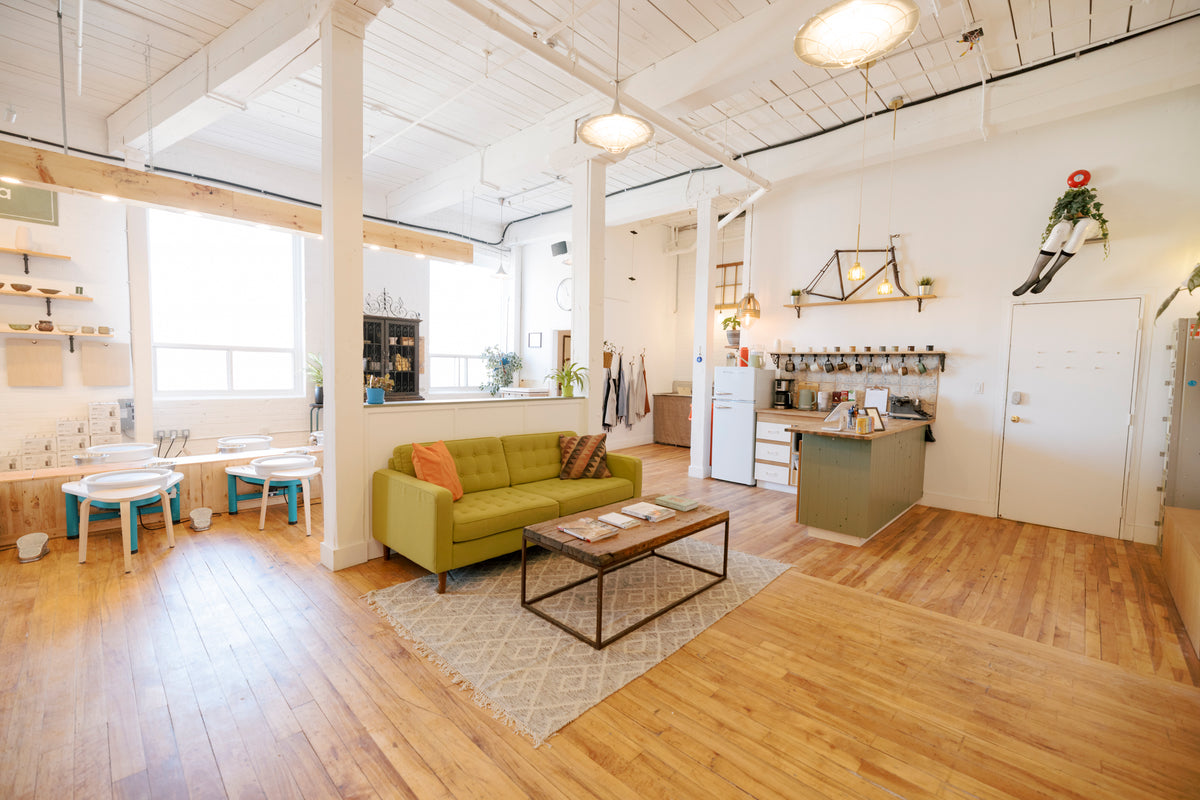 A bright, open loft-style living space with hardwood floors, exposed white ceiling beams, and a green sofa in the center. The room includes a small kitchen area, tables near the windows, shelving, and a patterned rug.