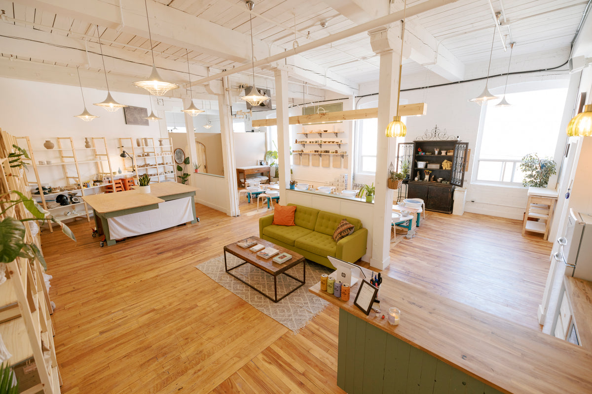 A bright, modern loft-style office or cafe interior with light wood floors, white columns, pendant lights, shelving, and a green sofa in the center of the room.