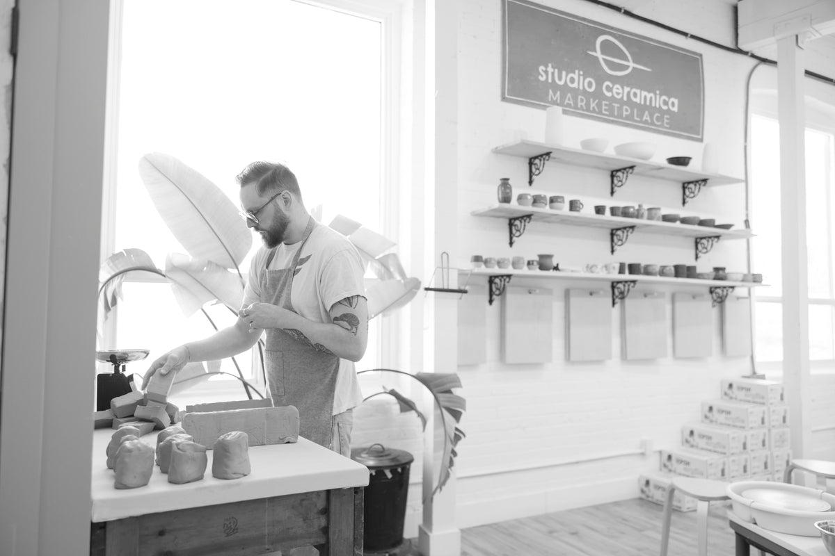 A black and white photograph of a ceramic artist working at a pottery wheel in a bright studio, with shelves of ceramic pieces and a sign reading 'studio ceramica MARKETPLACE' on the wall.