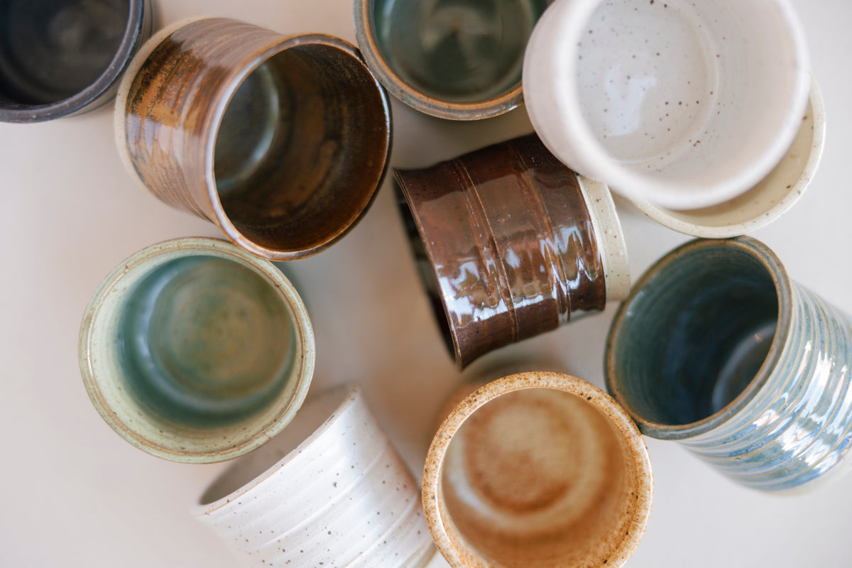 An overhead flat-lay arrangement of multiple handmade ceramic cups and bowls in various glazes, including brown, teal, blue, white, and speckled beige, on a light neutral background.