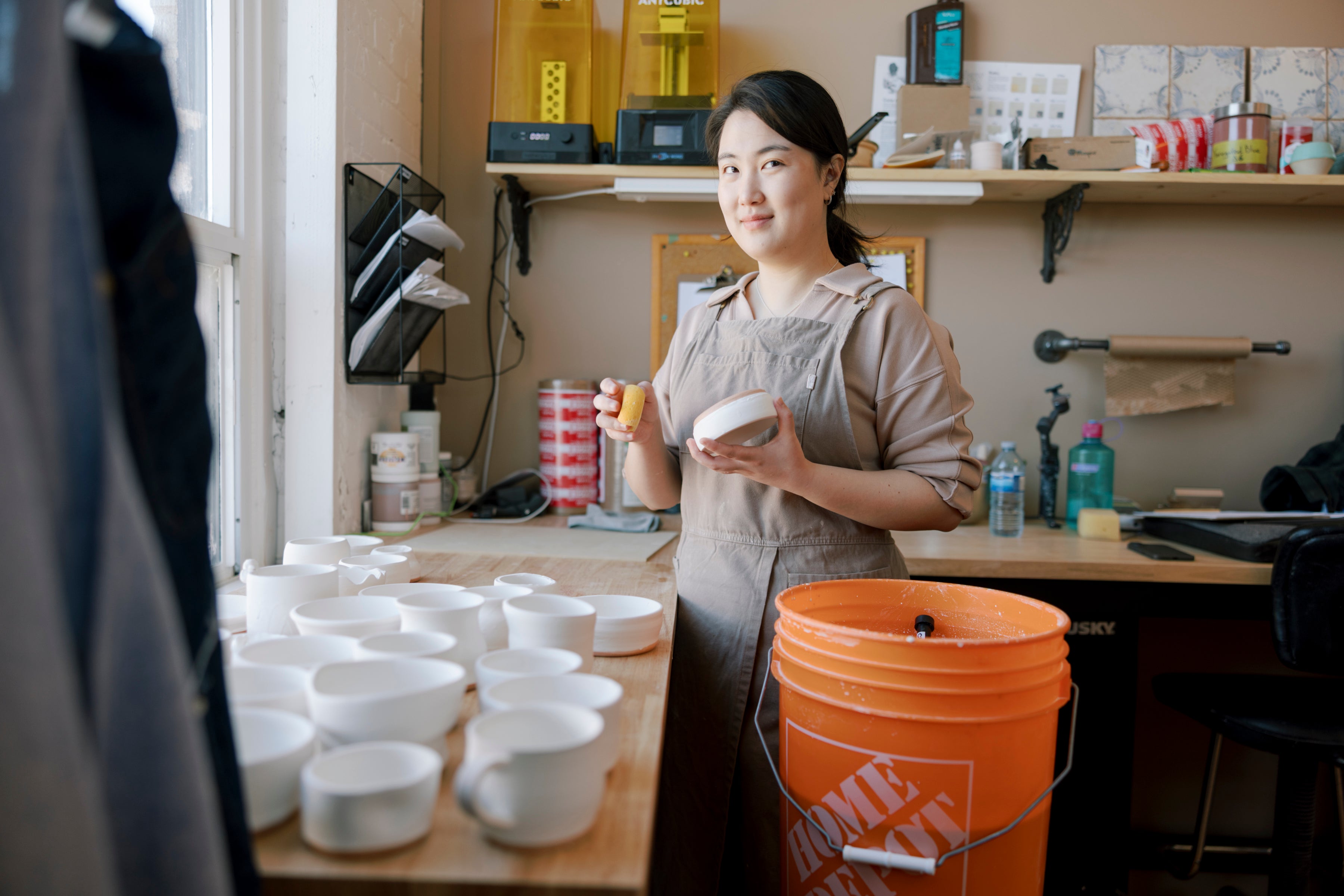A woman in a beige apron stands in a pottery studio holding a small white bowl, with many white ceramic bowls and cups arranged on a wooden table in the foreground and an orange bucket nearby.