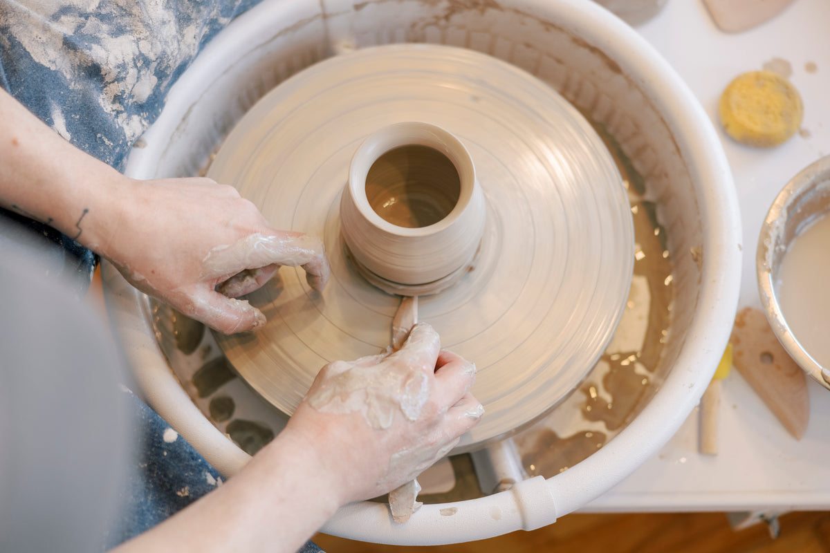 A close-up of a person's hands shaping a small clay pot on a spinning pottery wheel, with wet clay, tools, and a work surface visible.