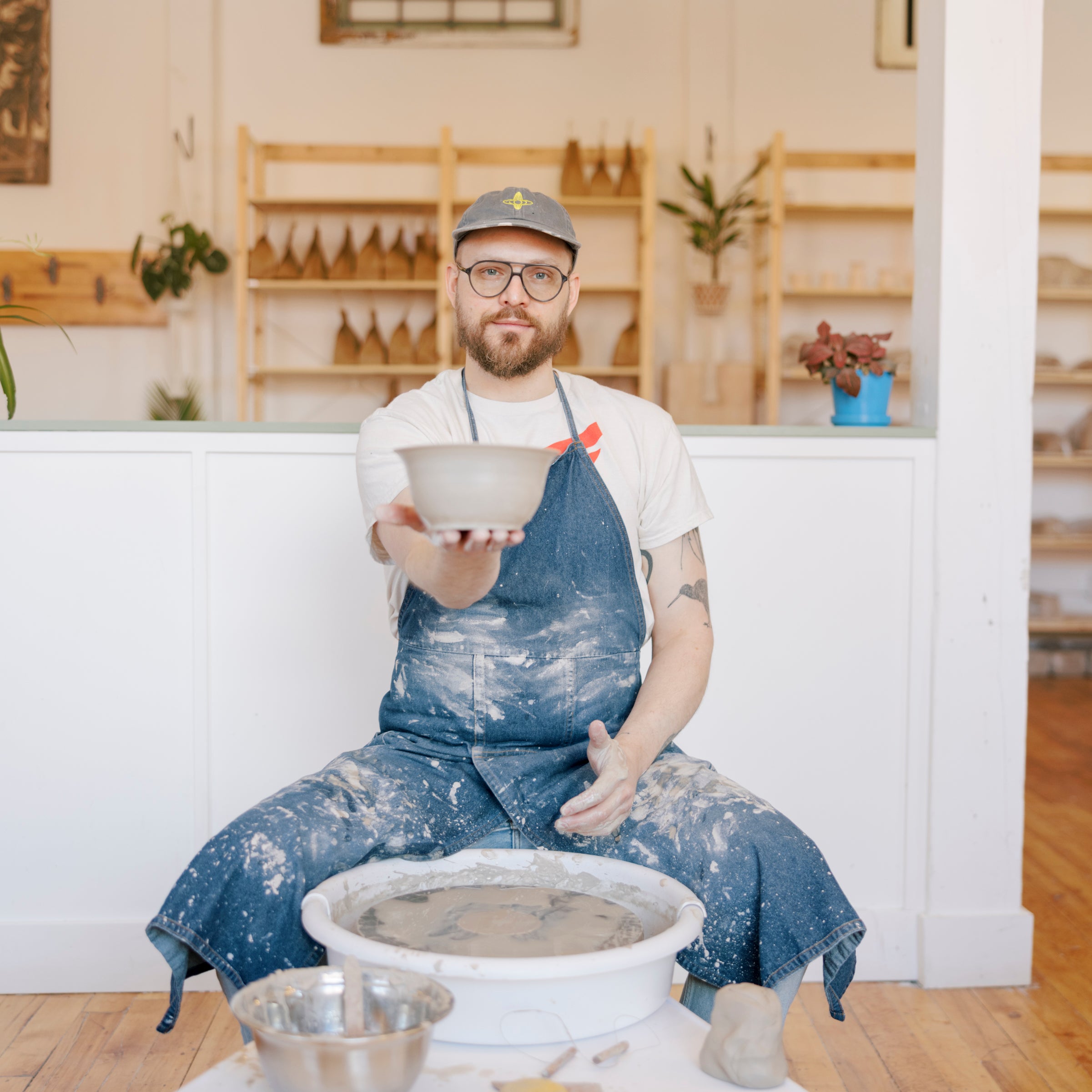 A man wearing a blue apron and cap sits in a pottery studio holding a small ceramic bowl, with a pottery wheel and clay bowls in front of him.