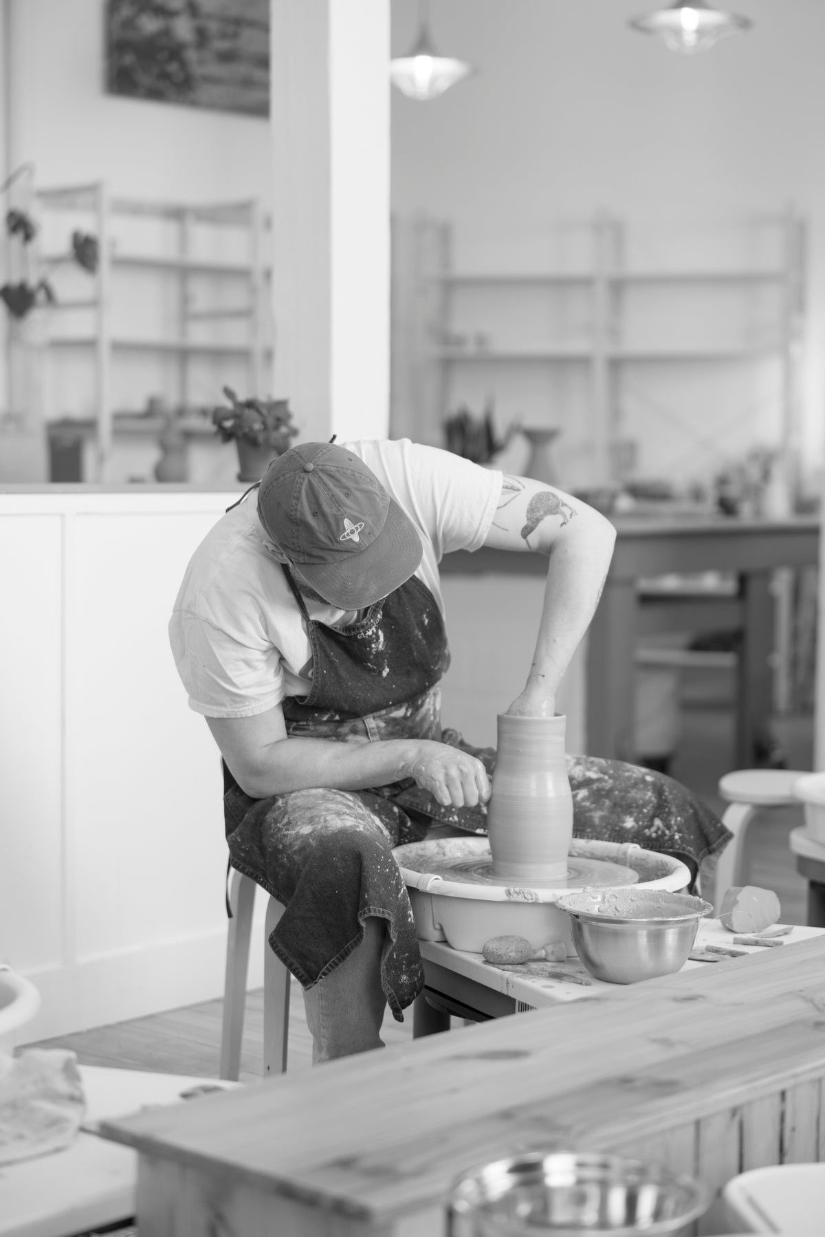 A black and white photograph of a potter shaping a clay vase on a pottery wheel in a studio.