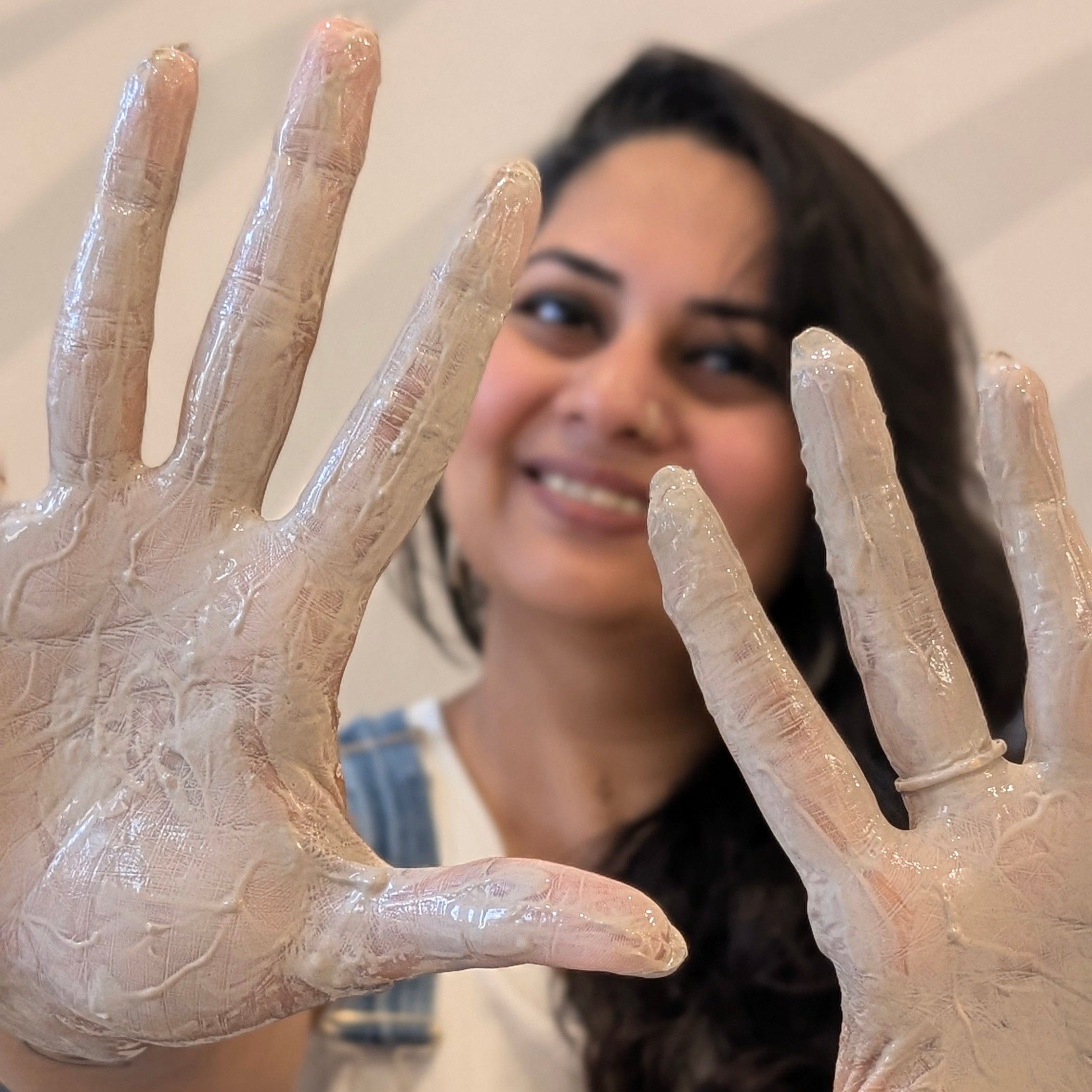 A woman with her face out of focus in the backgroud up her clay covered hands in focus. 