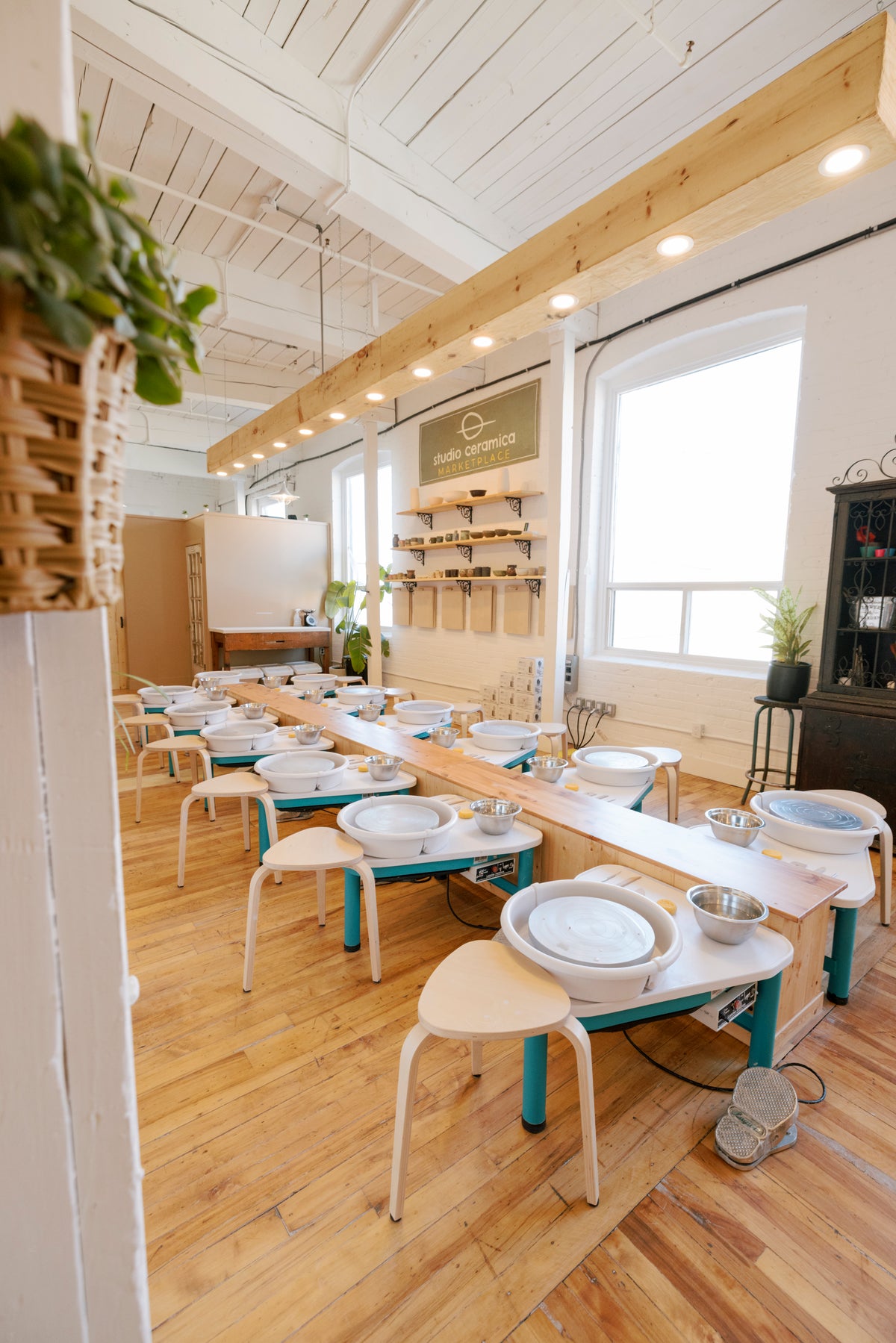 A beautiful pottery studio showing two rows of pottery wheels with wooden floors some shelves with pottery and a light beam above a center island. 