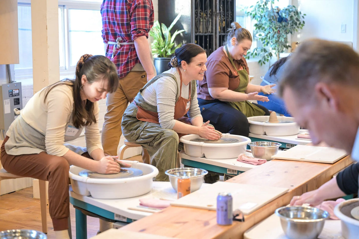 Several people seated at a pottery workshop table, shaping clay at white wheel basins in a bright indoor studio with windows and plants in the background.