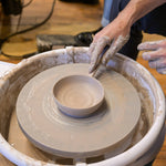 A close-up of a potter's wheel with a hand shaping a small clay bowl. A person's hands, covered in wet clay, are visible guiding the clay on the spinning wheel, set against a workshop background with a wooden floor.