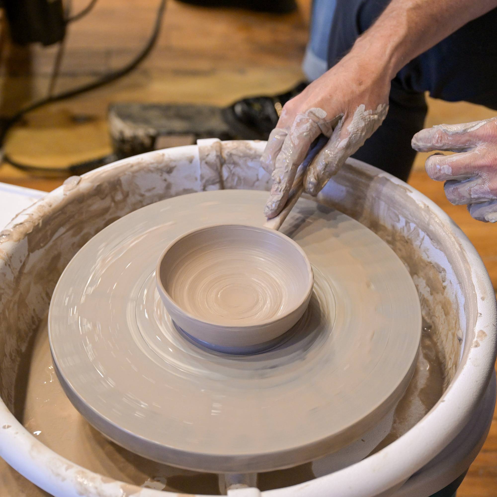 A close-up of a potter's wheel with a hand shaping a small clay bowl. A person's hands, covered in wet clay, are visible guiding the clay on the spinning wheel, set against a workshop background with a wooden floor.